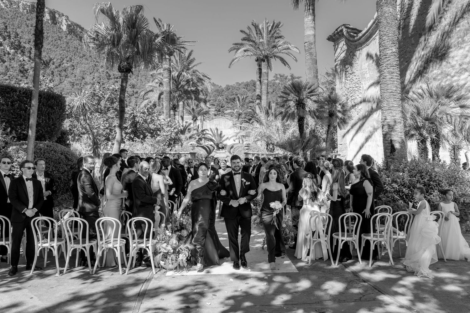 Black and white outdoor wedding ceremony with guests seated among palm trees and lush greenery.