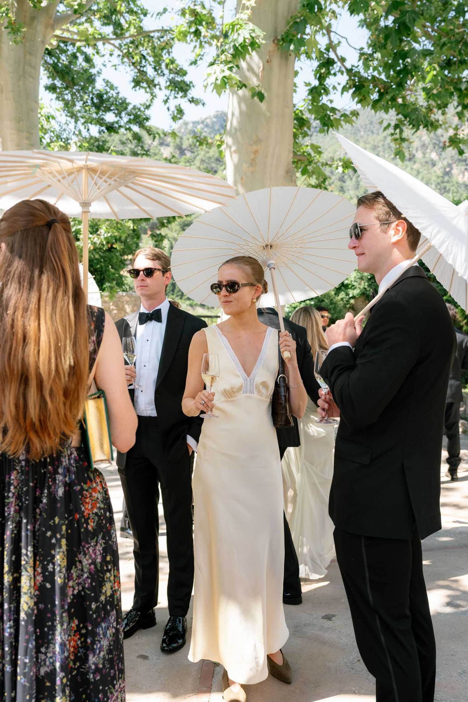 Elegant wedding guests in formal attire holding white parasols outdoors under sunny trees.
