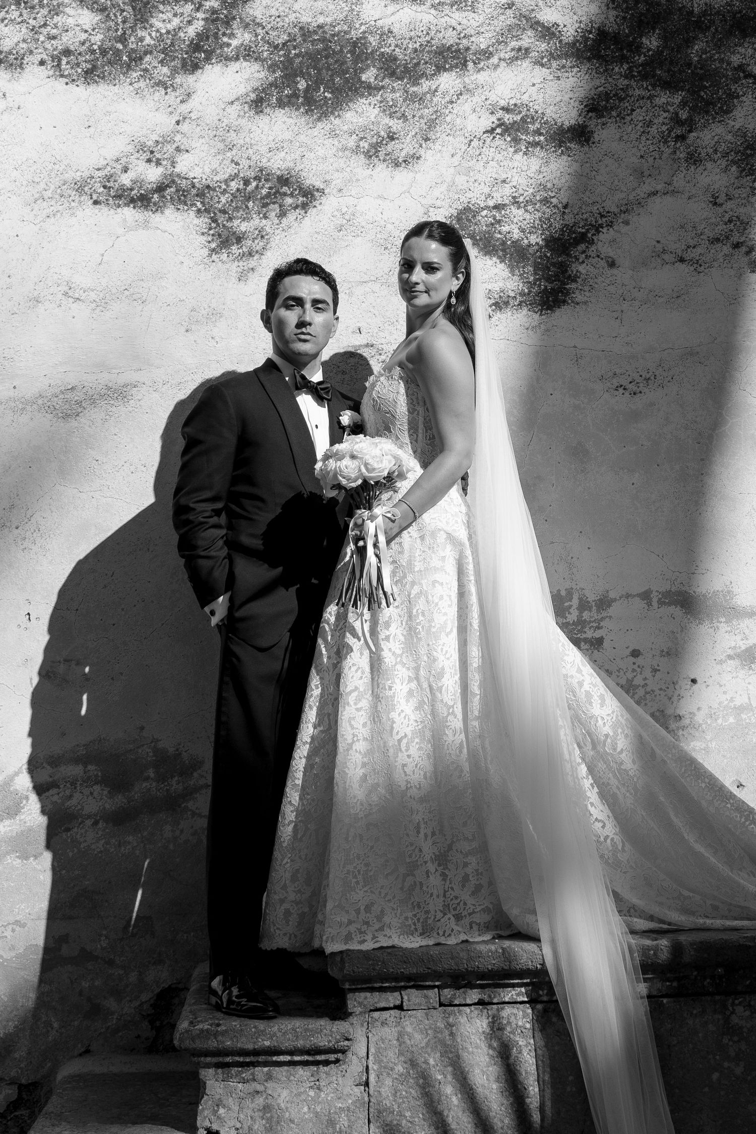 Black and white wedding photo of bride in lace gown with veil and groom in tuxedo posing against textured wall.