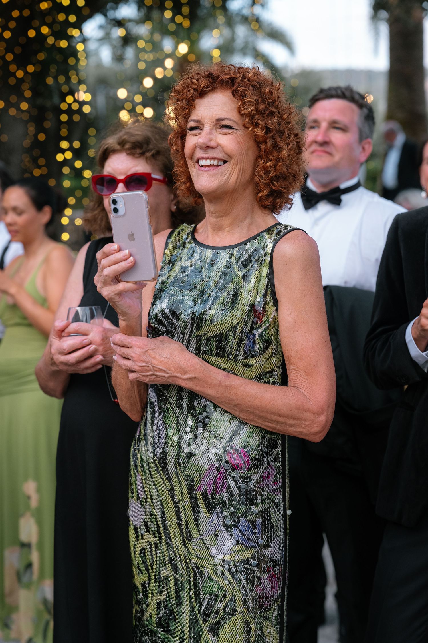Smiling woman in sequined floral dress holds phone at elegant outdoor evening event with guests in formal attire.
