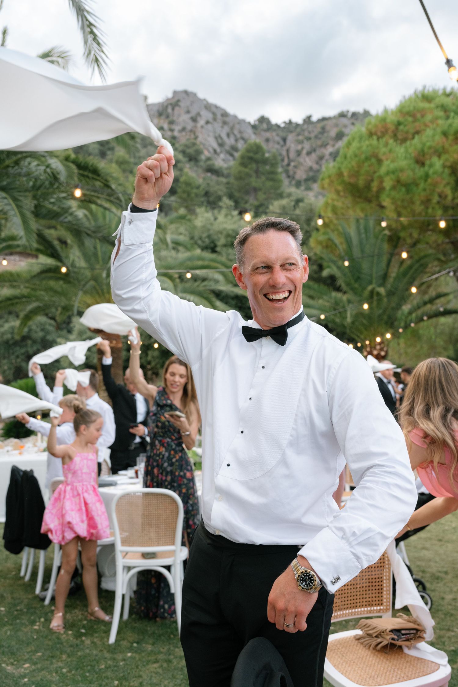 Man in white shirt and bow tie laughing and waving napkin at outdoor wedding reception with mountain backdrop.