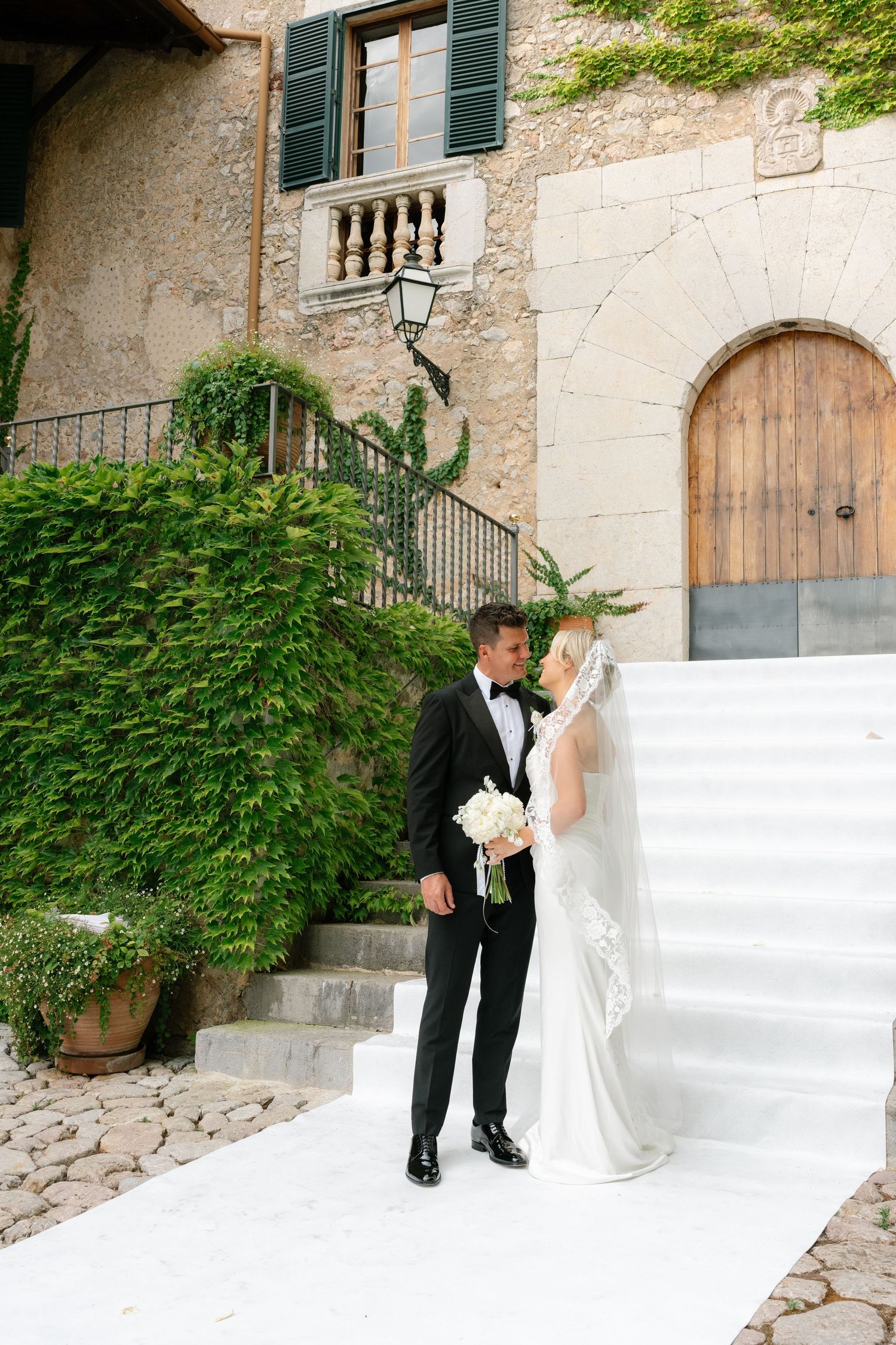 Bride and groom share a romantic moment on white steps outside a rustic stone villa with lush green ivy.