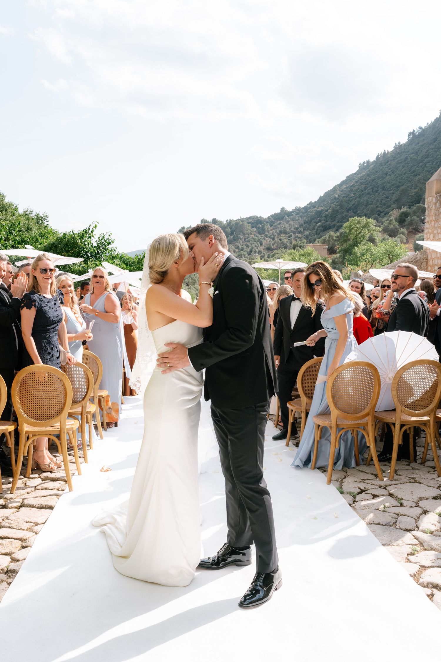 Bride and groom share first kiss at outdoor wedding ceremony with mountain backdrop and seated guests.