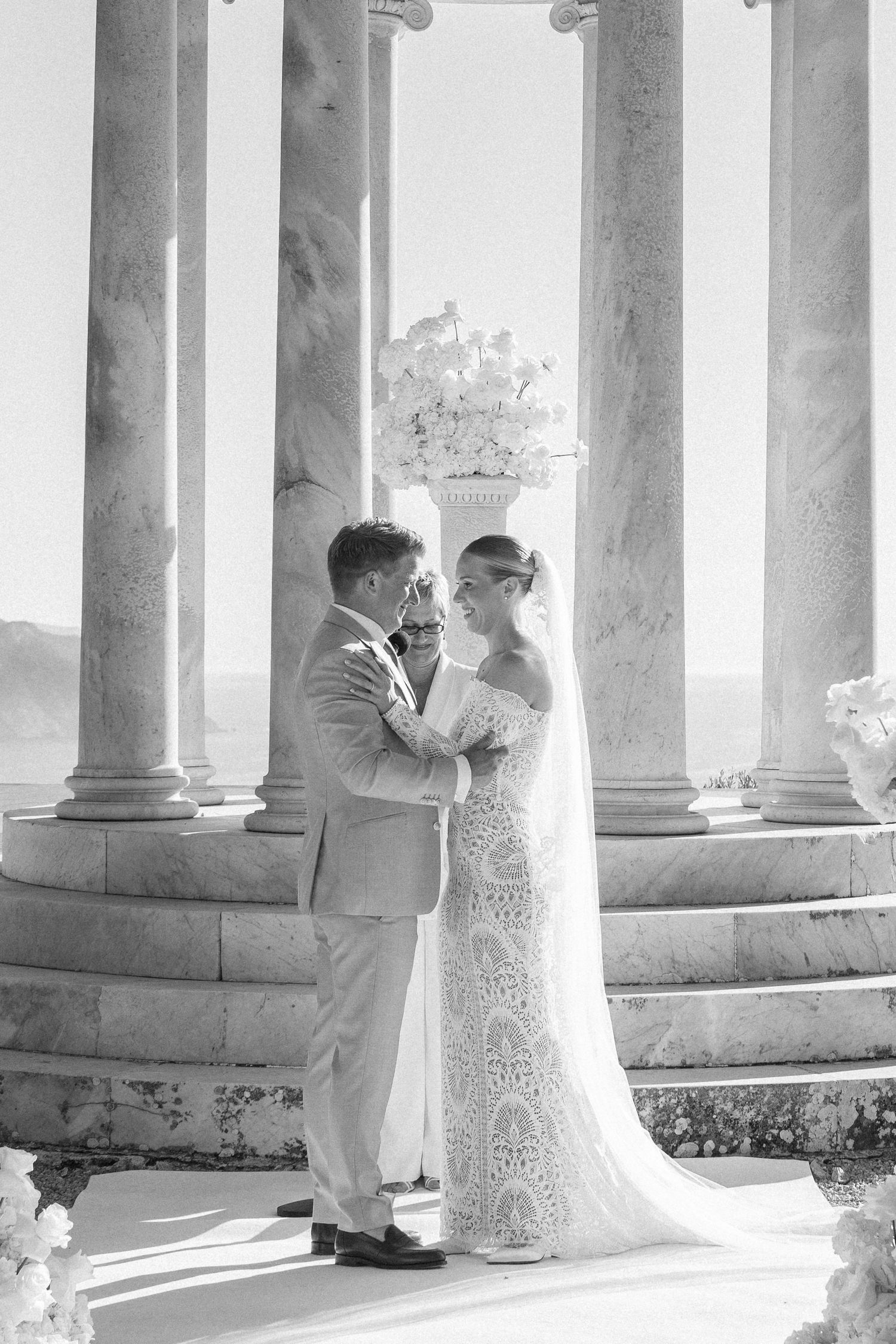 Wedding couple embracing between classical white columns in dramatic lighting.
