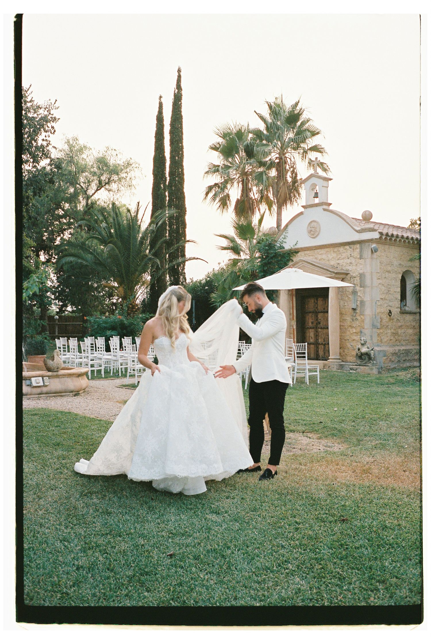 Wedding couple walking on grass with palm trees and stone building in background.