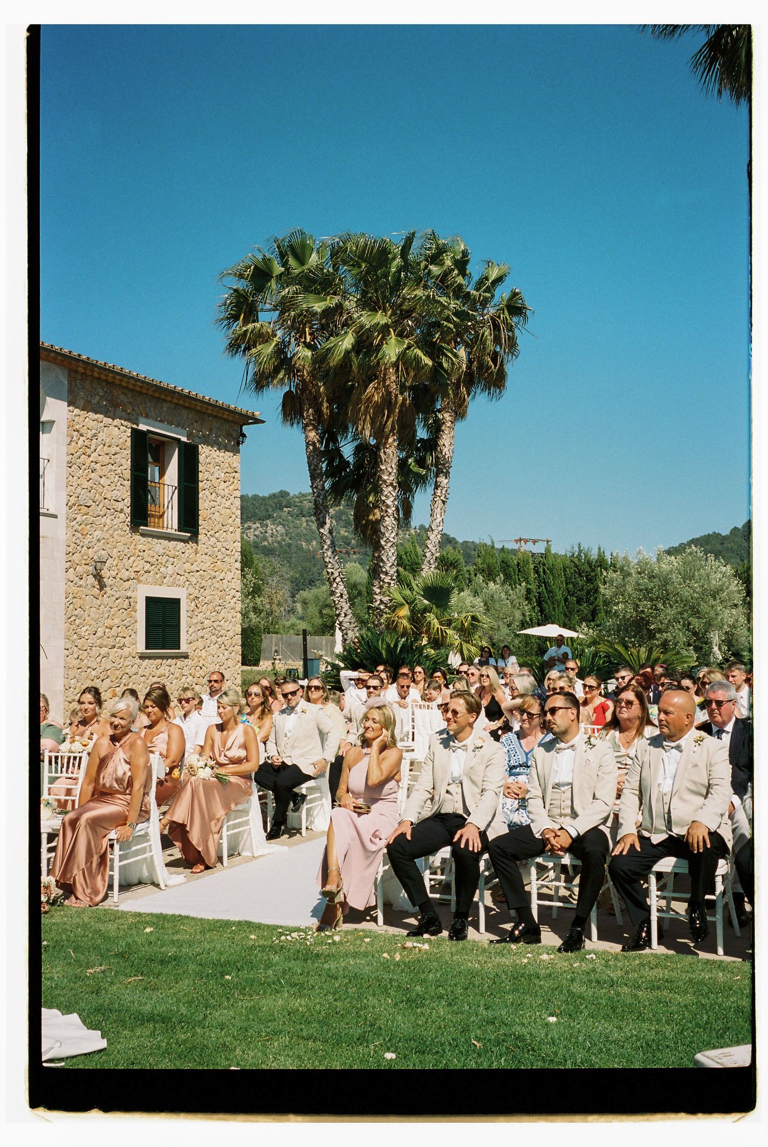 Wedding ceremony guests seated on a lawn with palm trees and stone buildings in view.