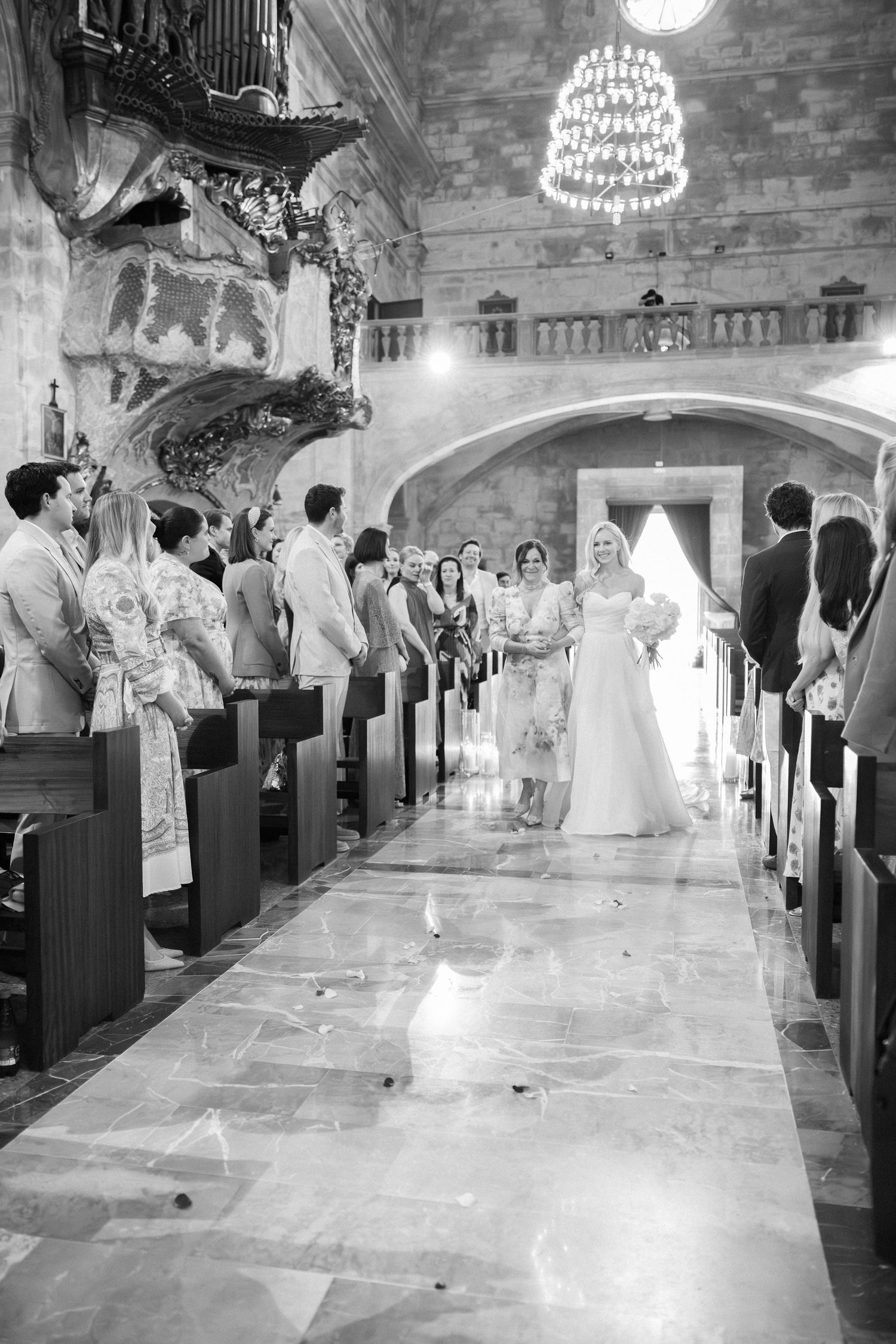 Black and white photo of a traditional church wedding ceremony with guests seated in wooden pews under chandeliers.