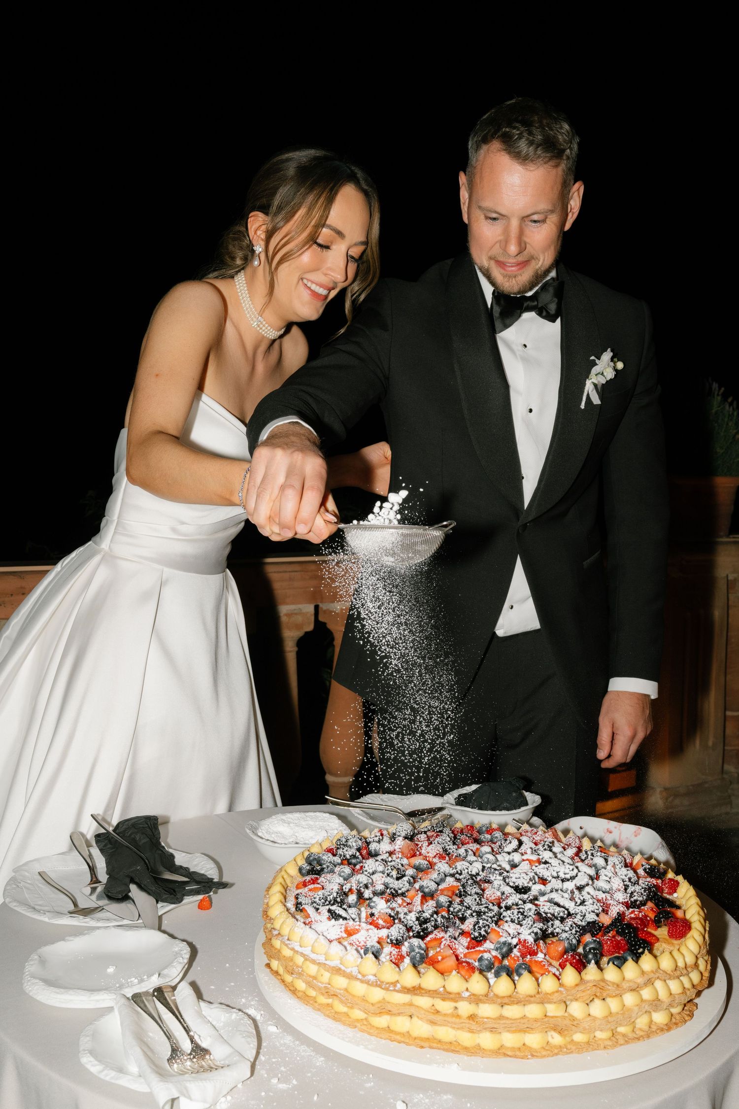 Newlyweds sprinkling powdered sugar on their multi-tiered wedding cake during reception.