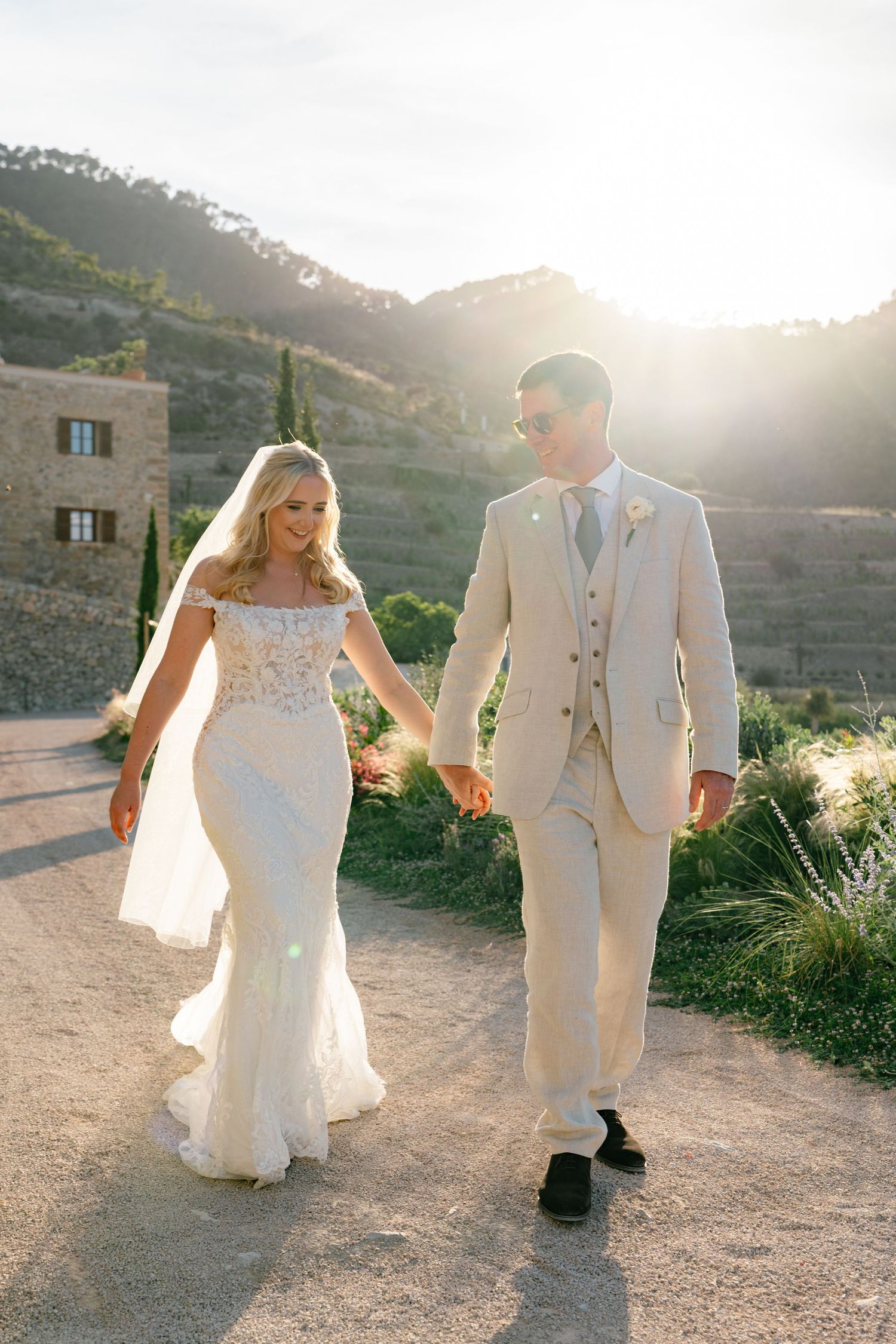 Newlyweds walking hand in hand on a sunlit mountain path in white attire.