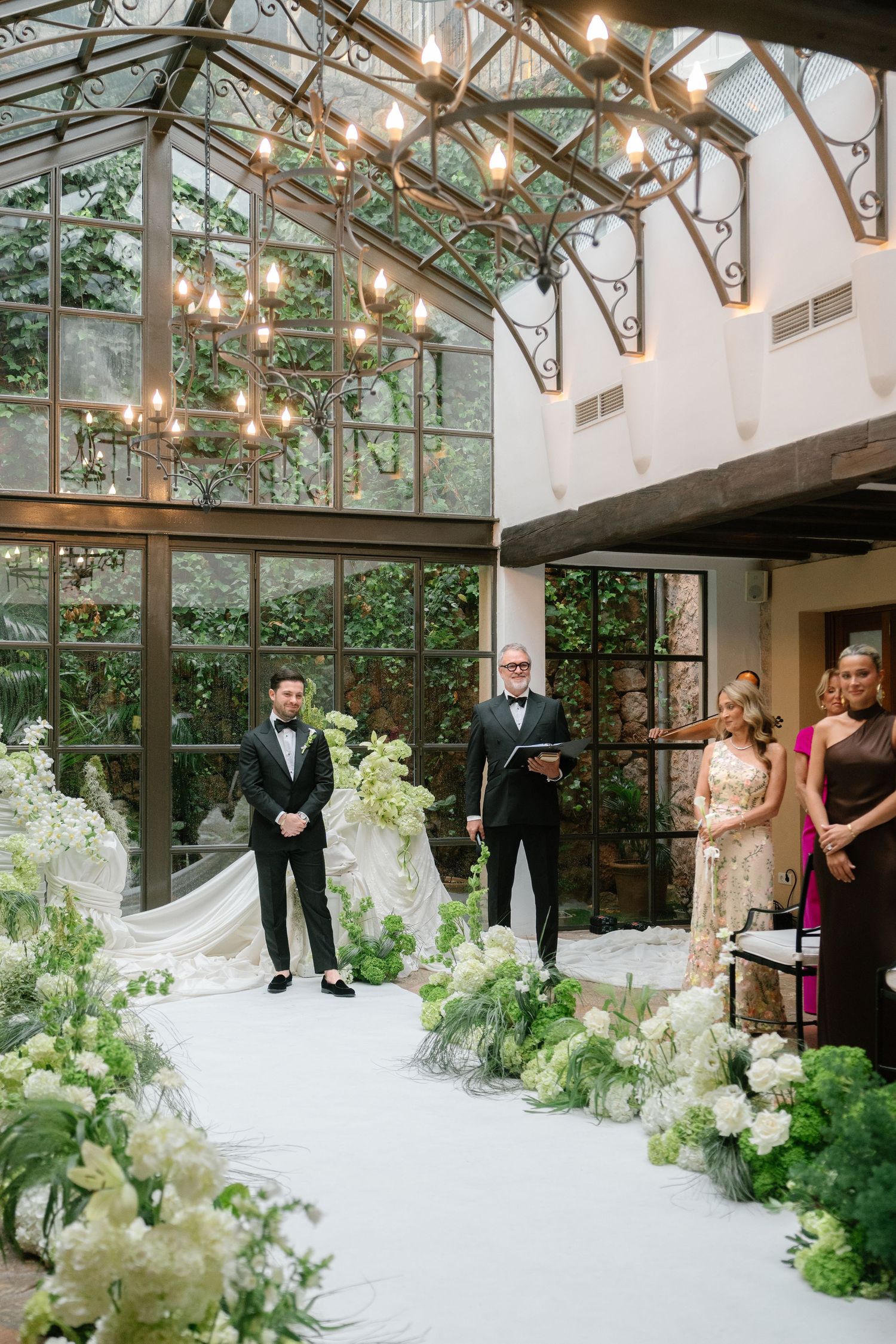 Wedding party standing in greenhouse venue with lush white floral arrangements and natural lighting.