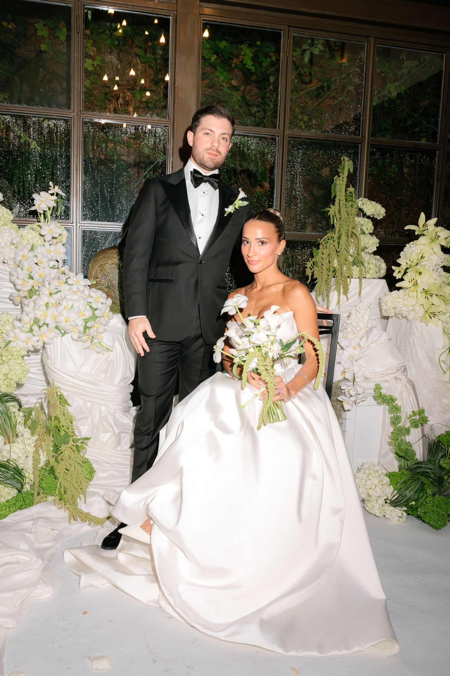 A bride and groom pose together in a romantic wedding setting with white floral decorations and elegant lighting.