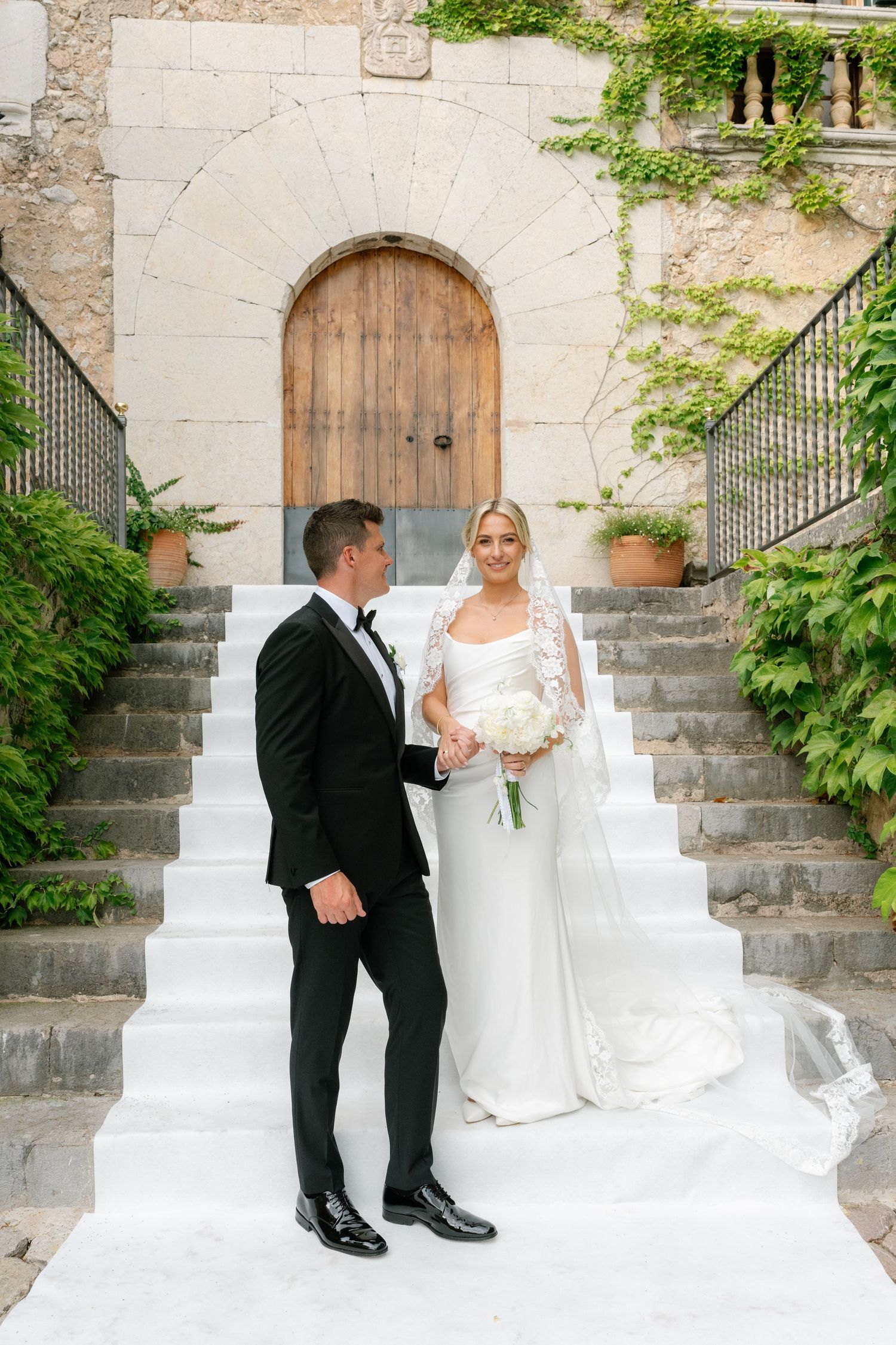 Bride and groom stand on white steps before a rustic stone building with wooden door and lush greenery.