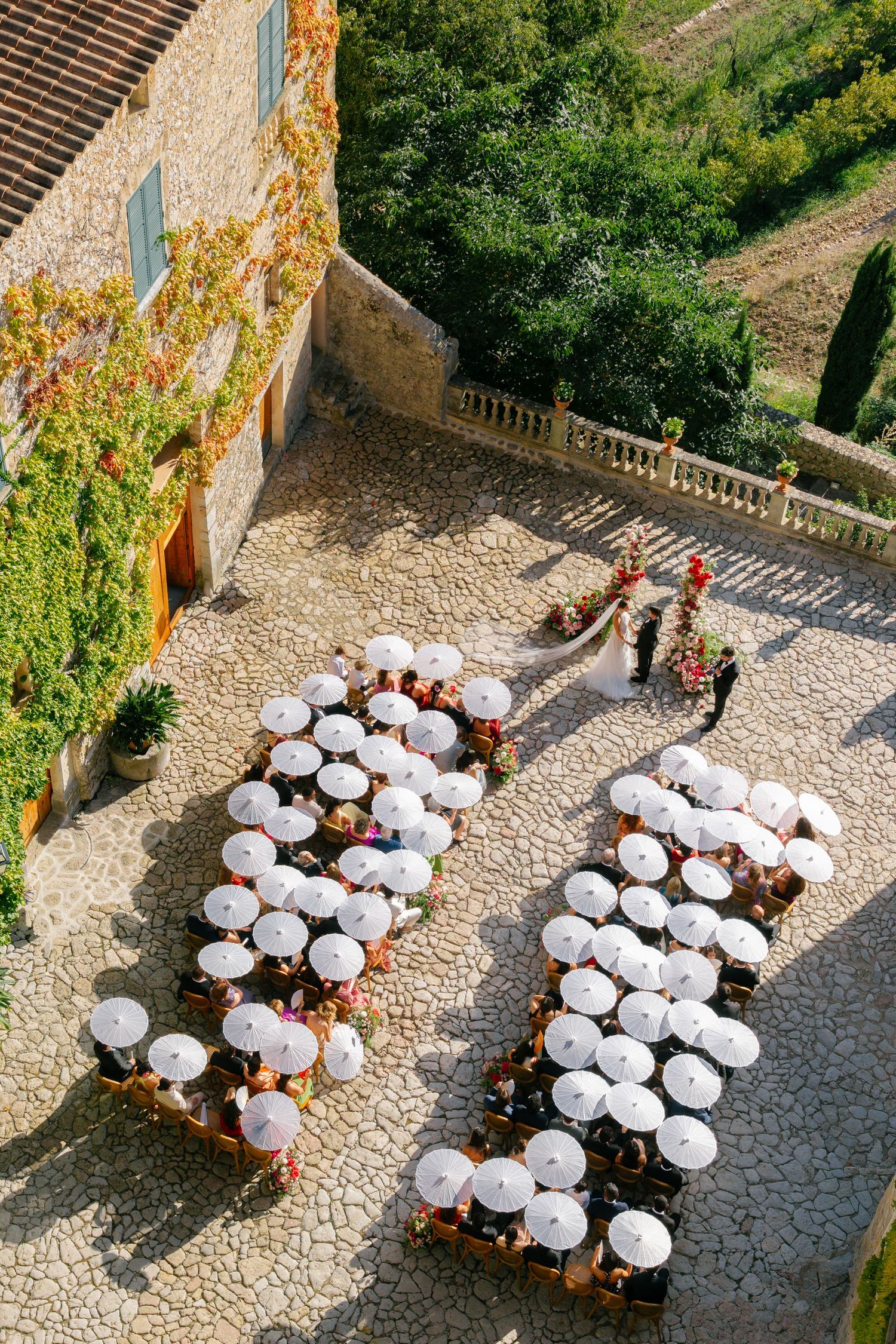 Aerial view of an outdoor wedding ceremony with guests seated under white parasols on a cobblestone courtyard at Finca Comassema