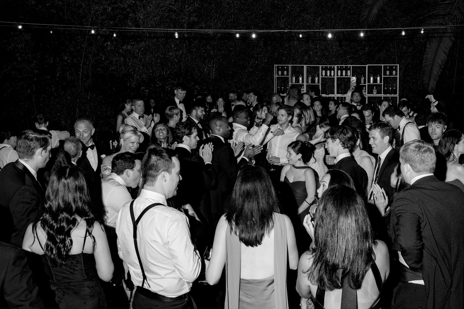 Black and white photo of a lively wedding reception dance floor filled with elegantly dressed guests celebrating.