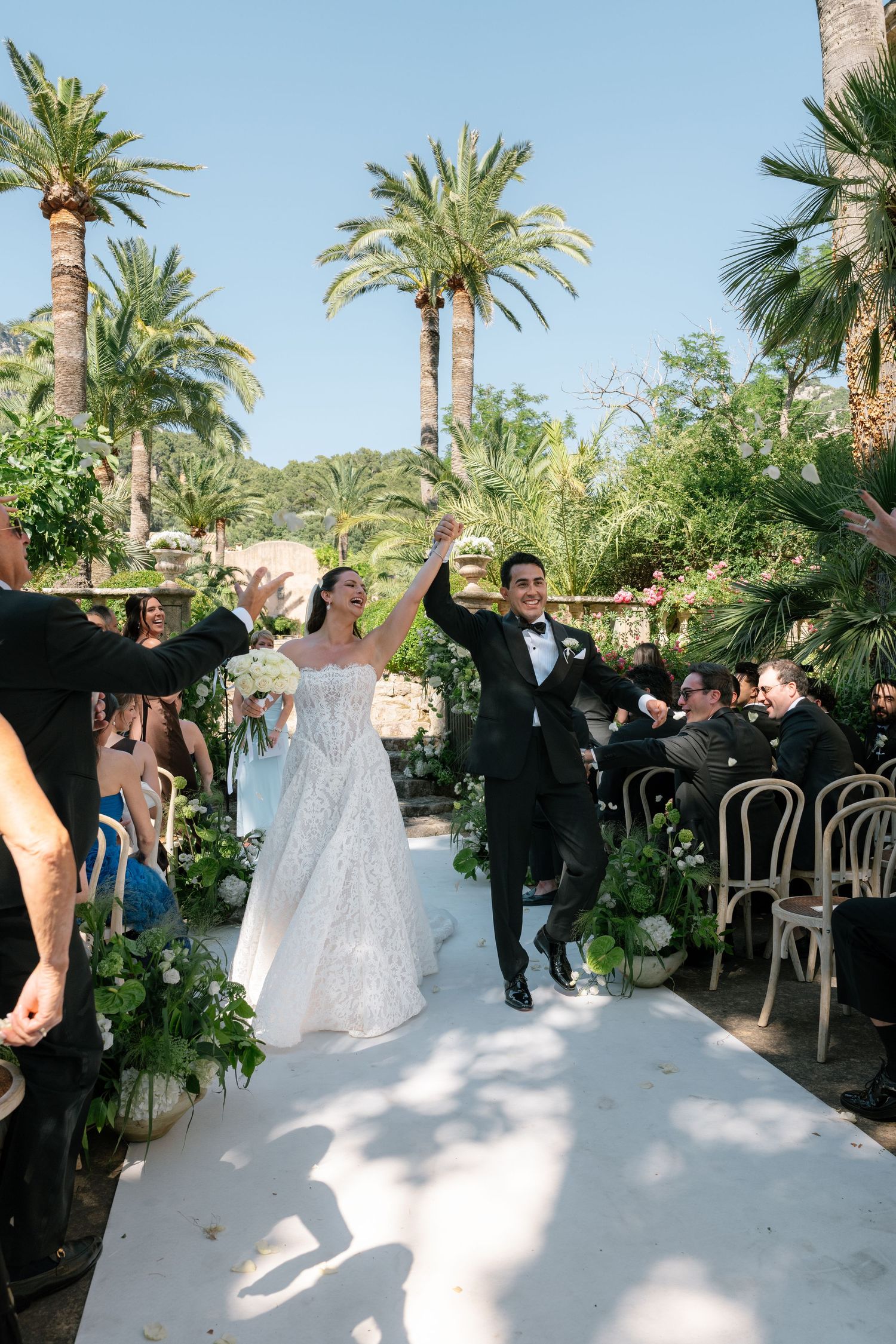 Bride and groom celebrate joyfully walking down outdoor aisle lined with greenery and palm trees on sunny wedding day.