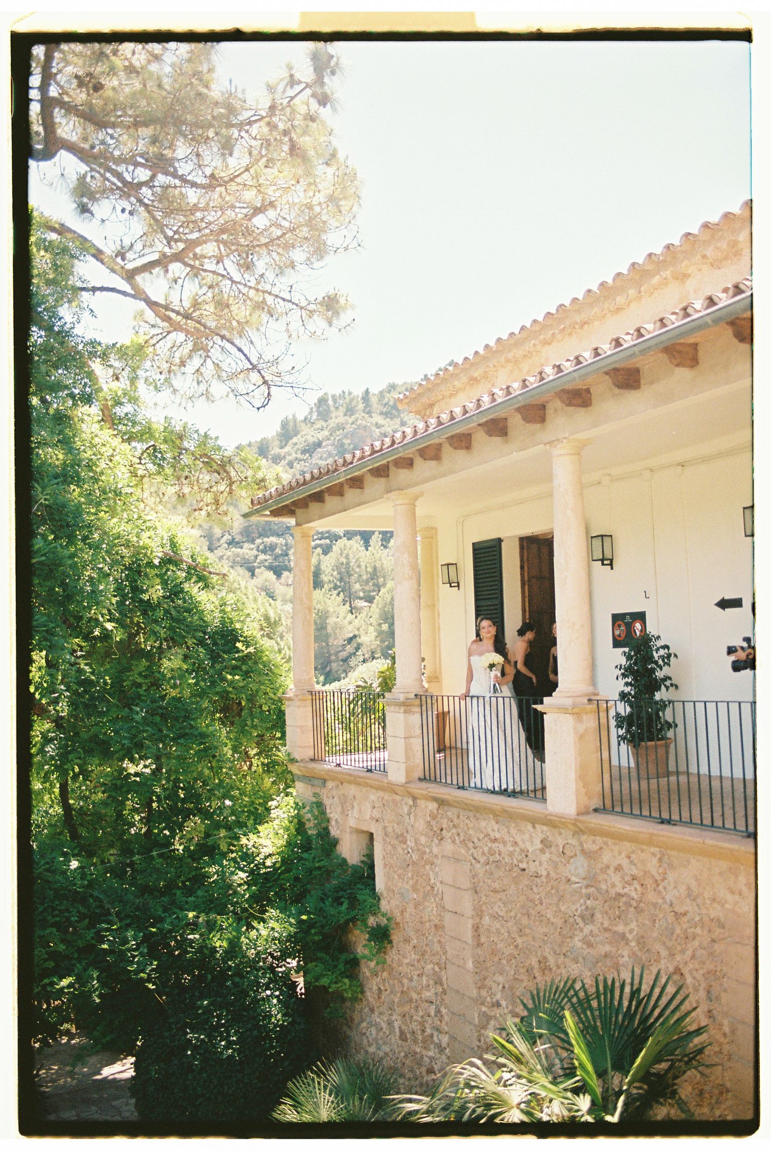 Elegant Mediterranean villa with columned porch surrounded by lush greenery, featuring a couple on the terrace on a sunny day.