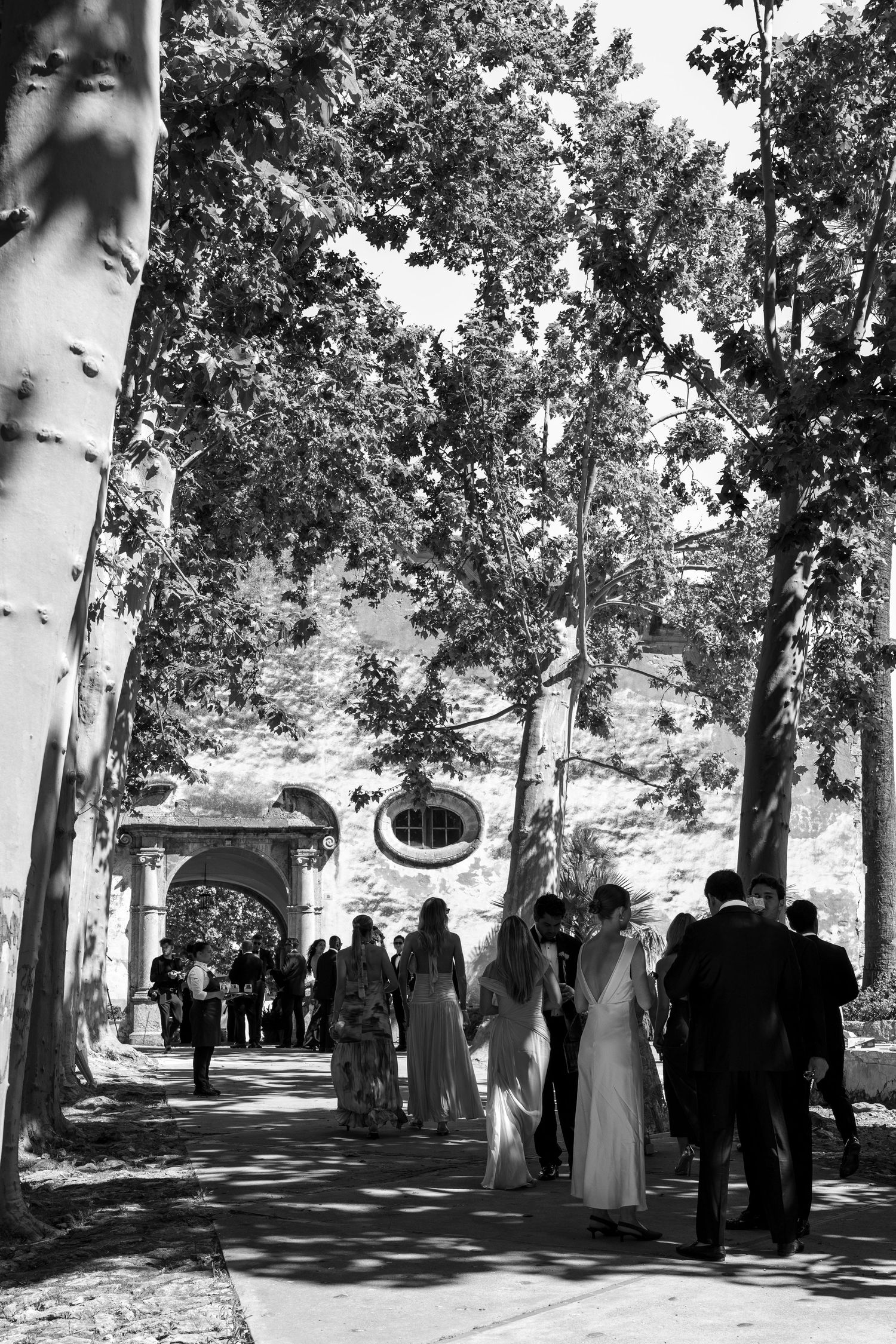 Black and white photo of wedding guests walking along a tree-lined path toward a stone chapel.