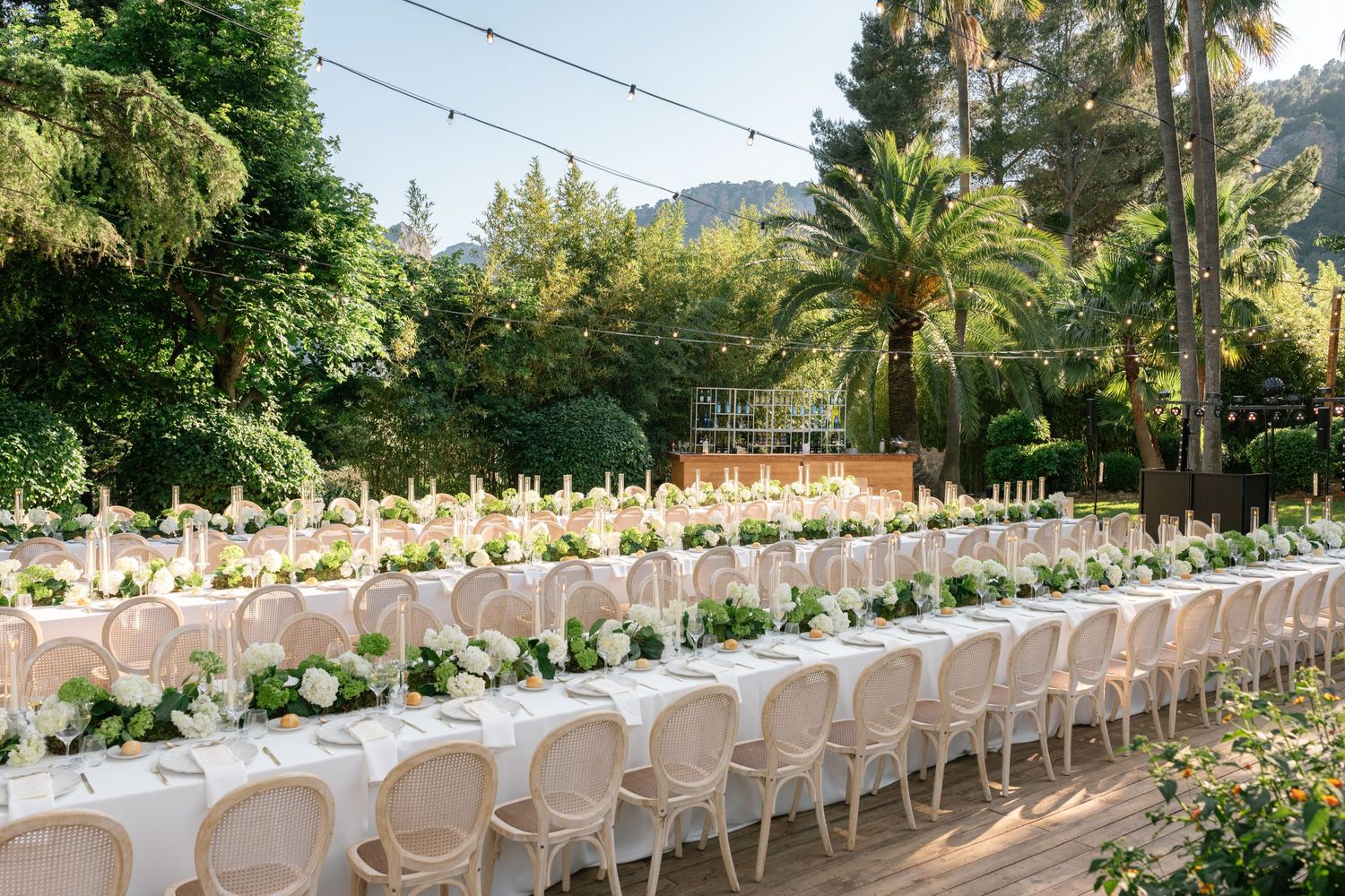 Elegant outdoor wedding reception with long white tables, floral centerpieces, and string lights among lush green trees.