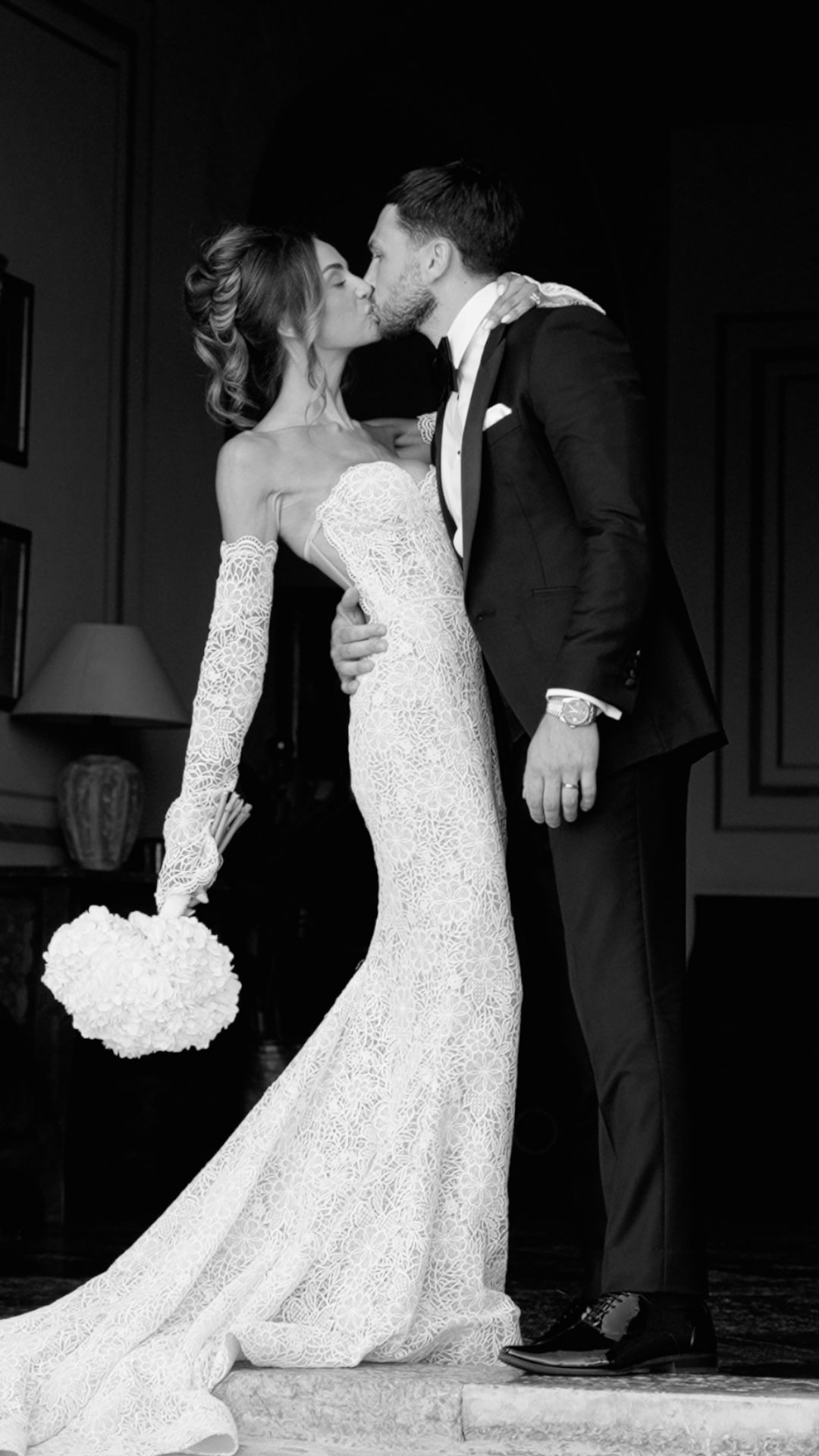 Black and white wedding photo of bride in lace gown with bouquet kissing groom in tuxedo.