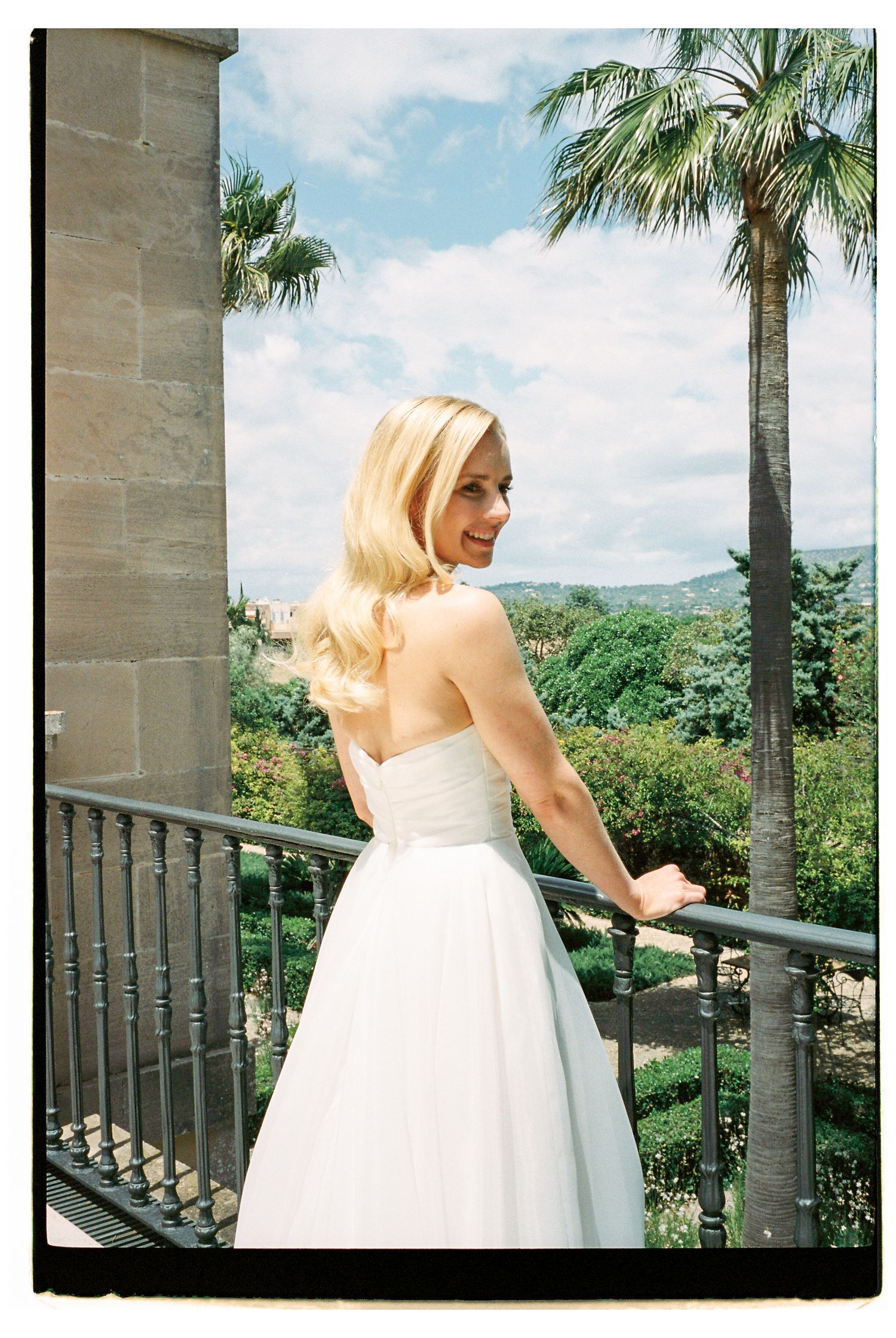 Bride in a white gown poses on a balcony with palm trees and blue sky in the background.