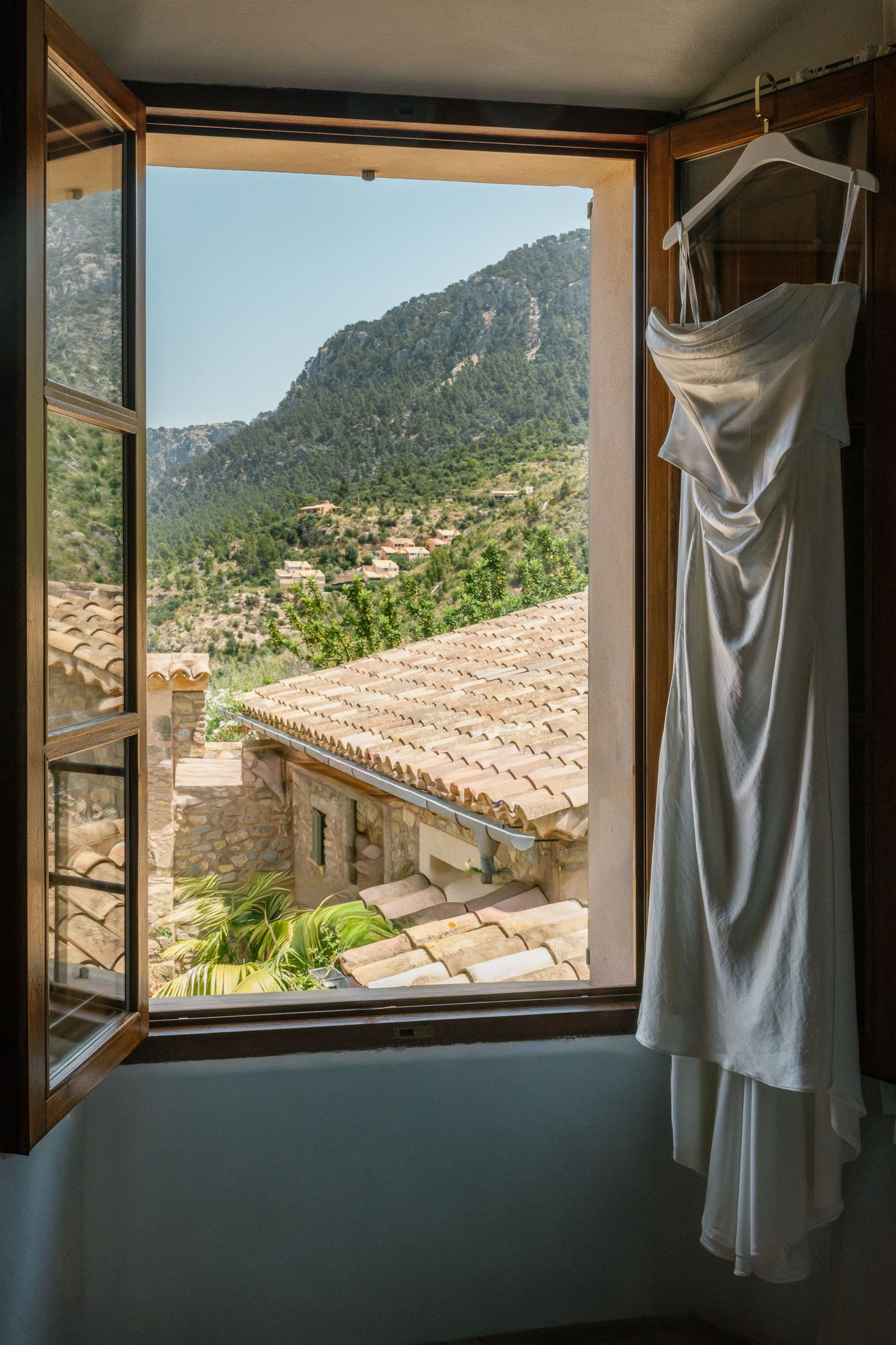 White wedding dress hanging by a window overlooking Mediterranean terracotta roofs and mountain scenery.