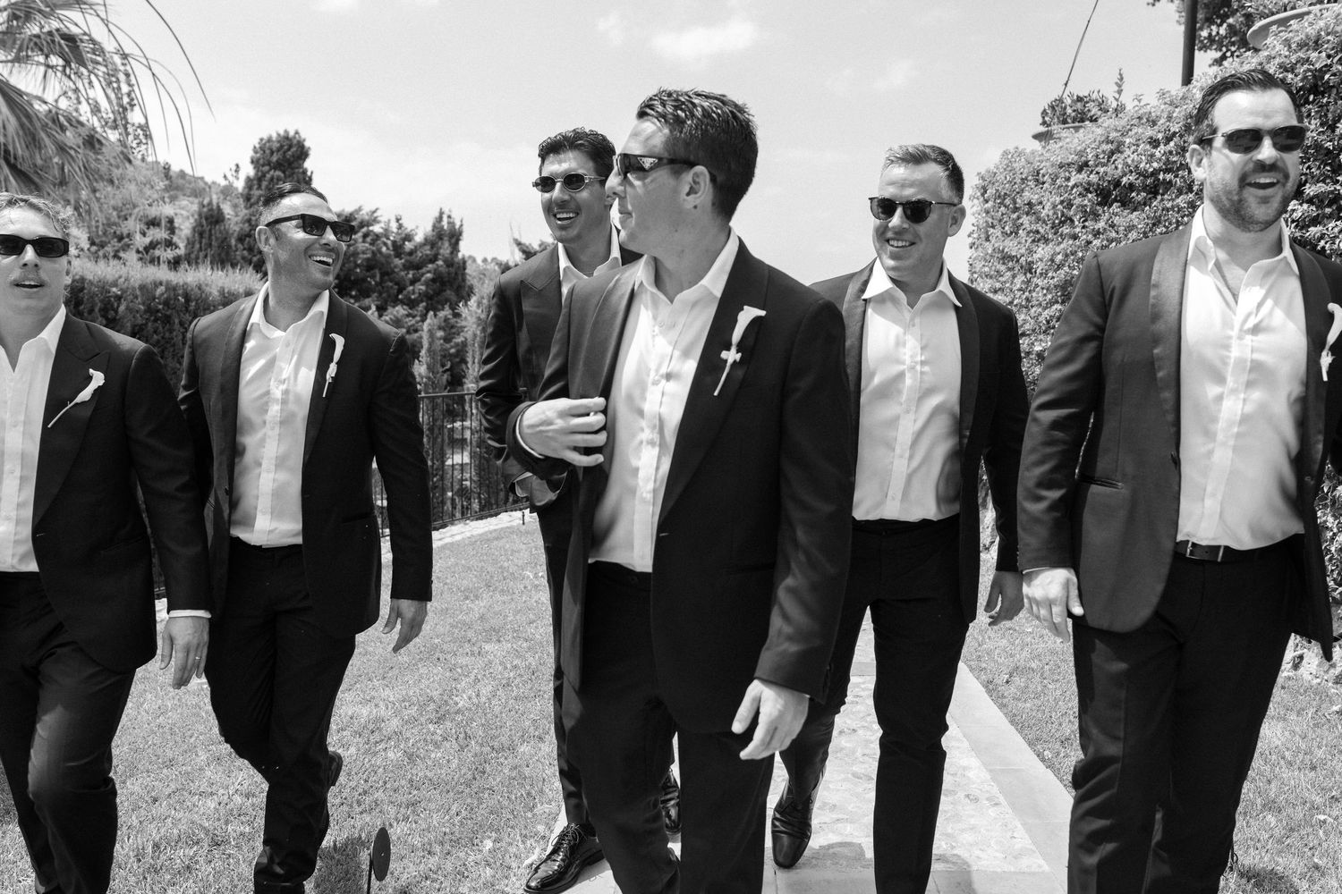 A group of groomsmen in formal black and white attire walk together outdoors while smiling and laughing.