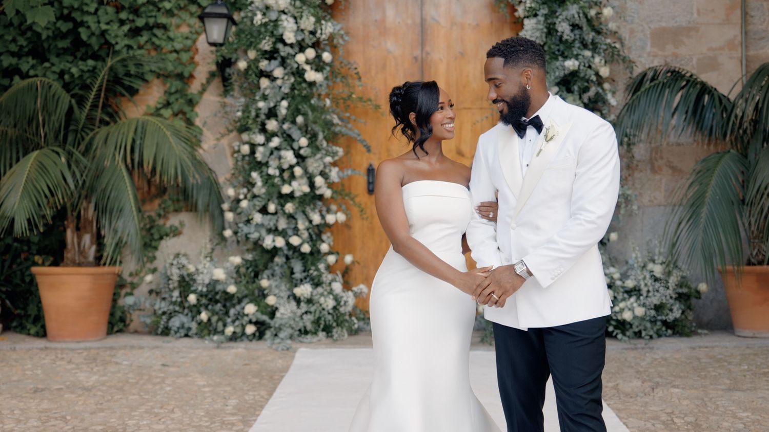 A couple shares intimate moments during a wedding photo session against a rustic stone wall with white floral decor.