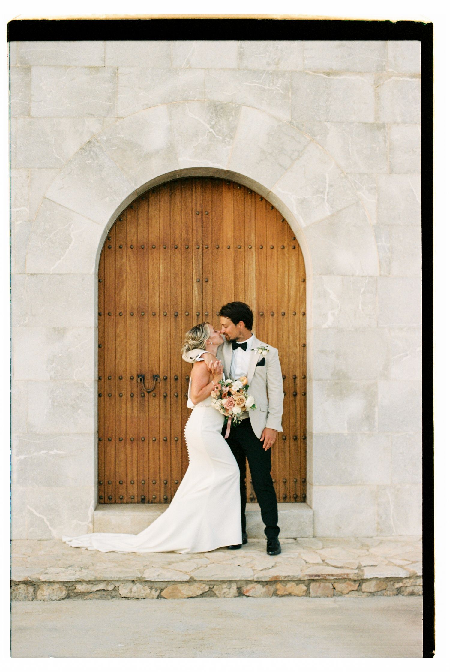 A romantic moment captured in front of wooden arched doors against a stone wall.