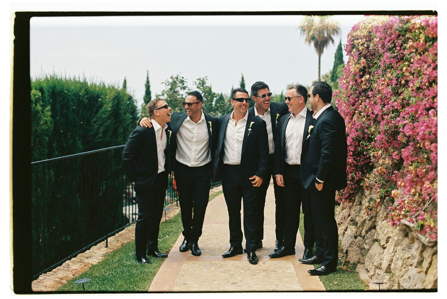 Groomsmen in formal attire gather on a Mediterranean pathway lined with pink bougainvillea.