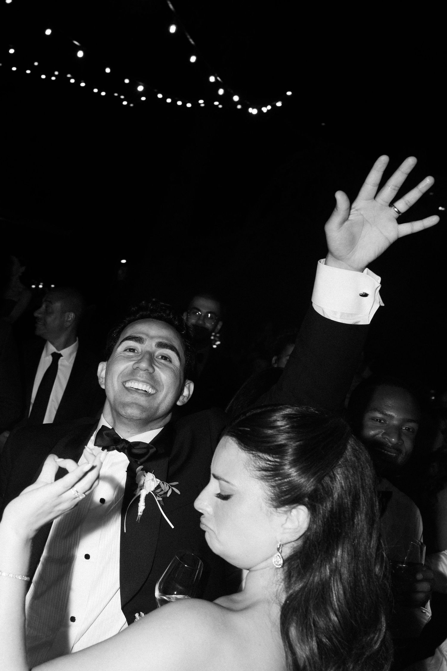 Black and white photo of a smiling groom in a tuxedo and bride dancing joyfully at their wedding reception.