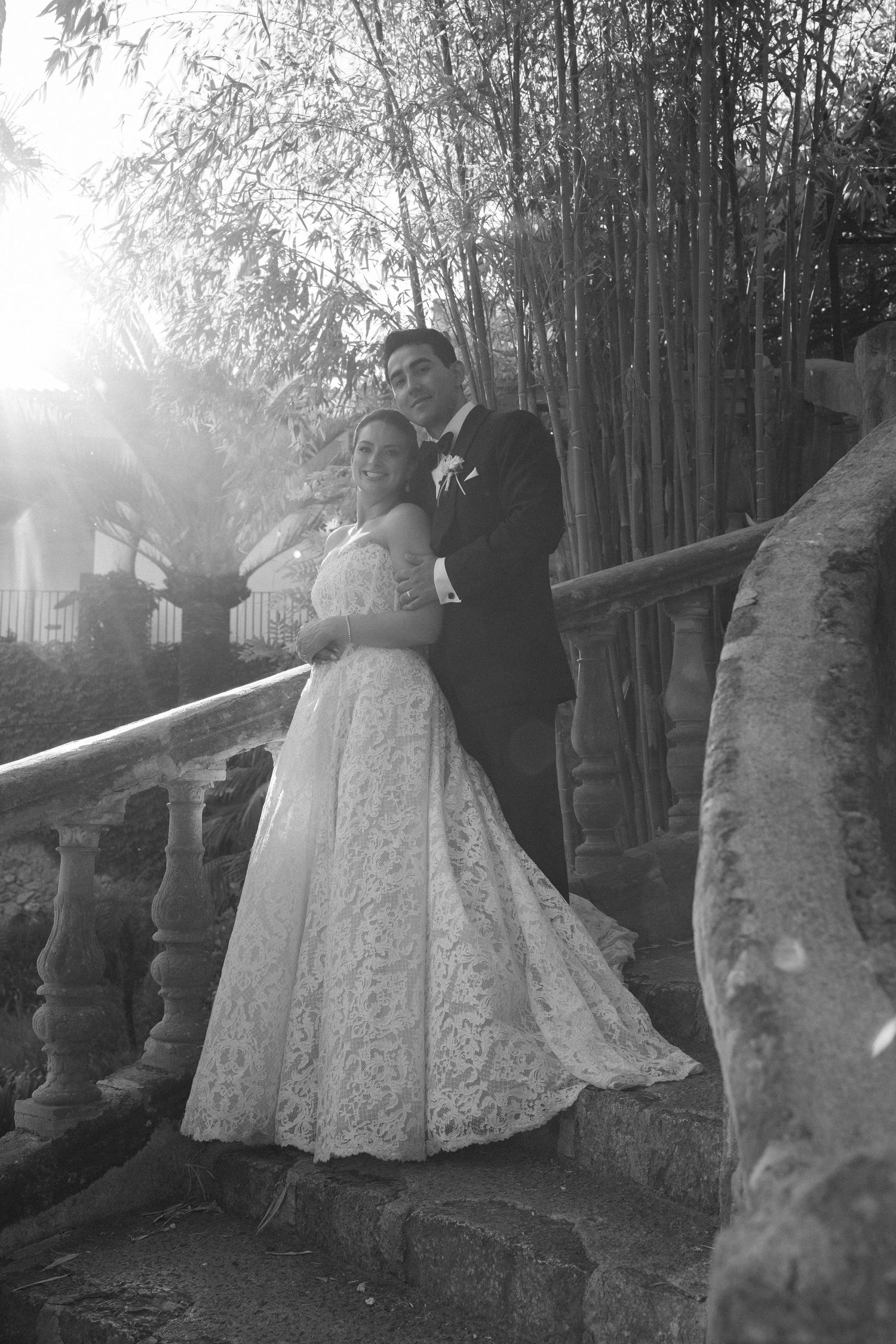 Black and white wedding photo of bride in lace gown and groom in tuxedo embracing on stone steps near bamboo.
