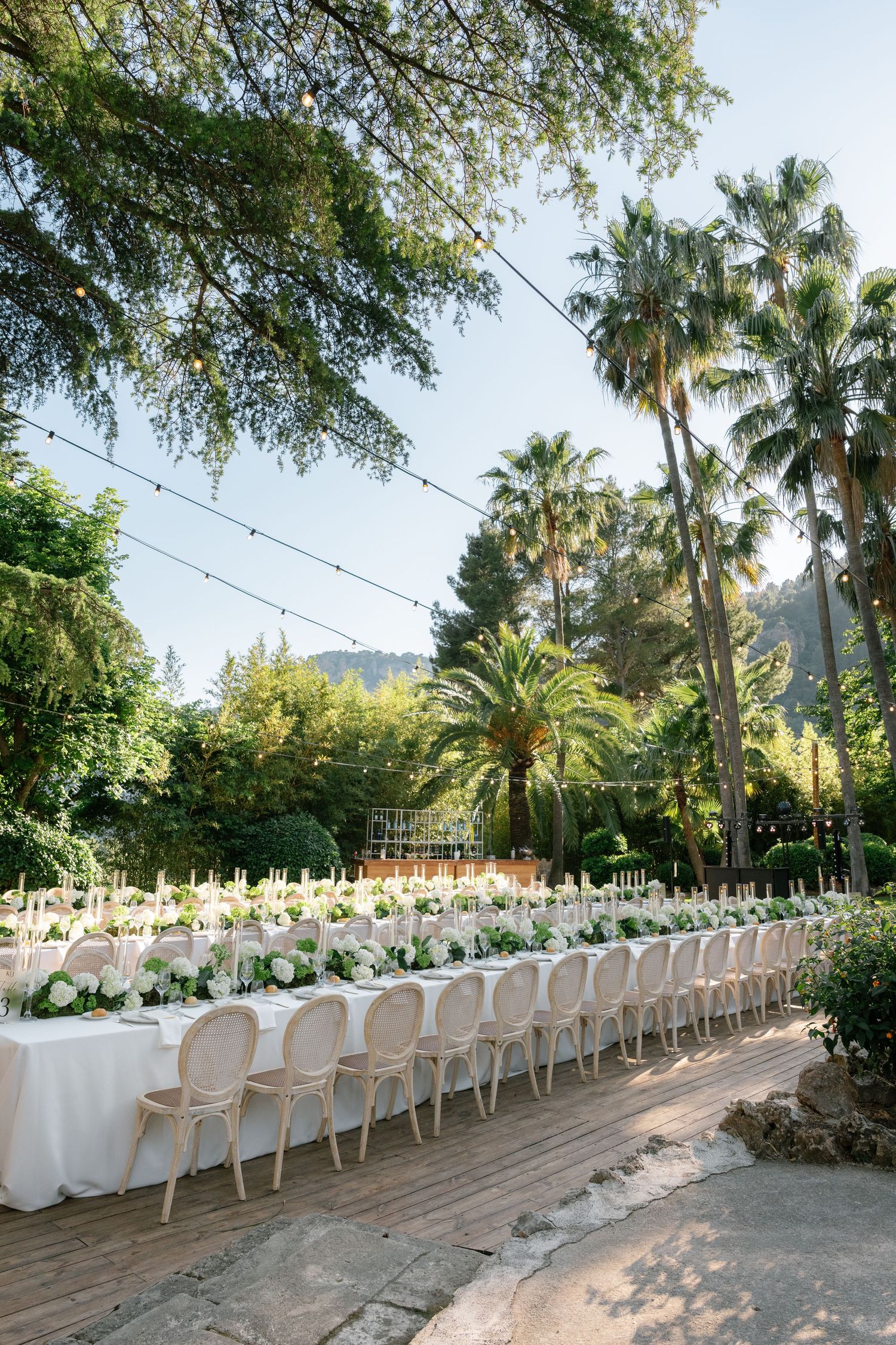 Elegant outdoor wedding reception with long white-draped tables, ghost chairs, floral centerpieces, string lights, and lush palm trees.