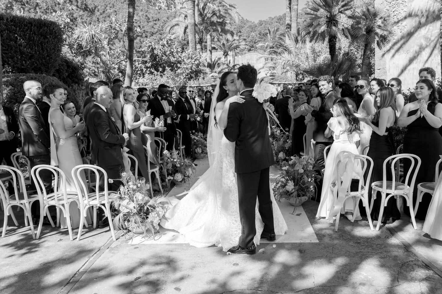 Black and white photo of a couple sharing their first kiss at an outdoor wedding ceremony surrounded by guests.