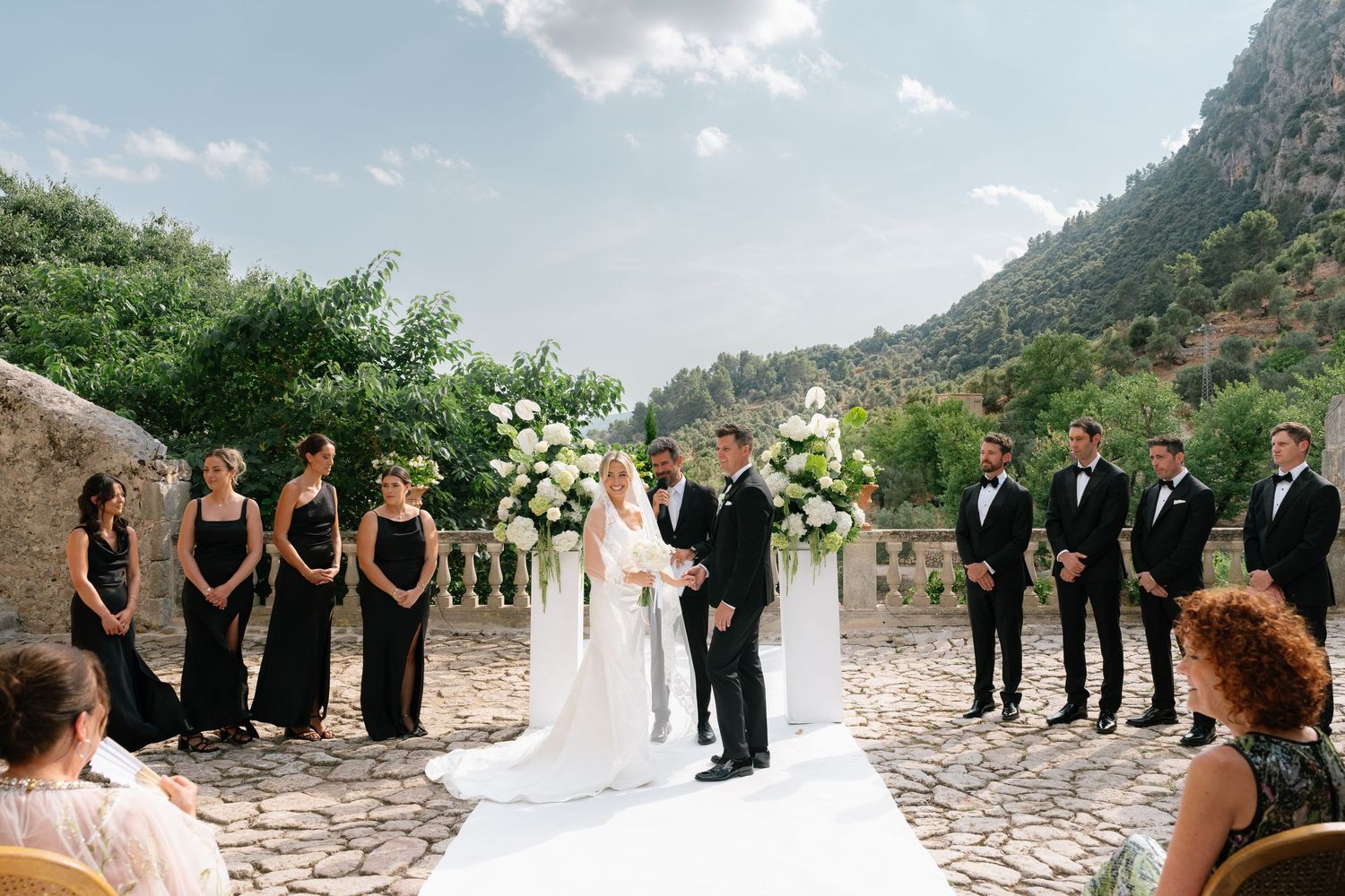 Outdoor wedding ceremony with bride and groom at altar, bridesmaids in black dresses, groomsmen in tuxedos, mountain backdrop.