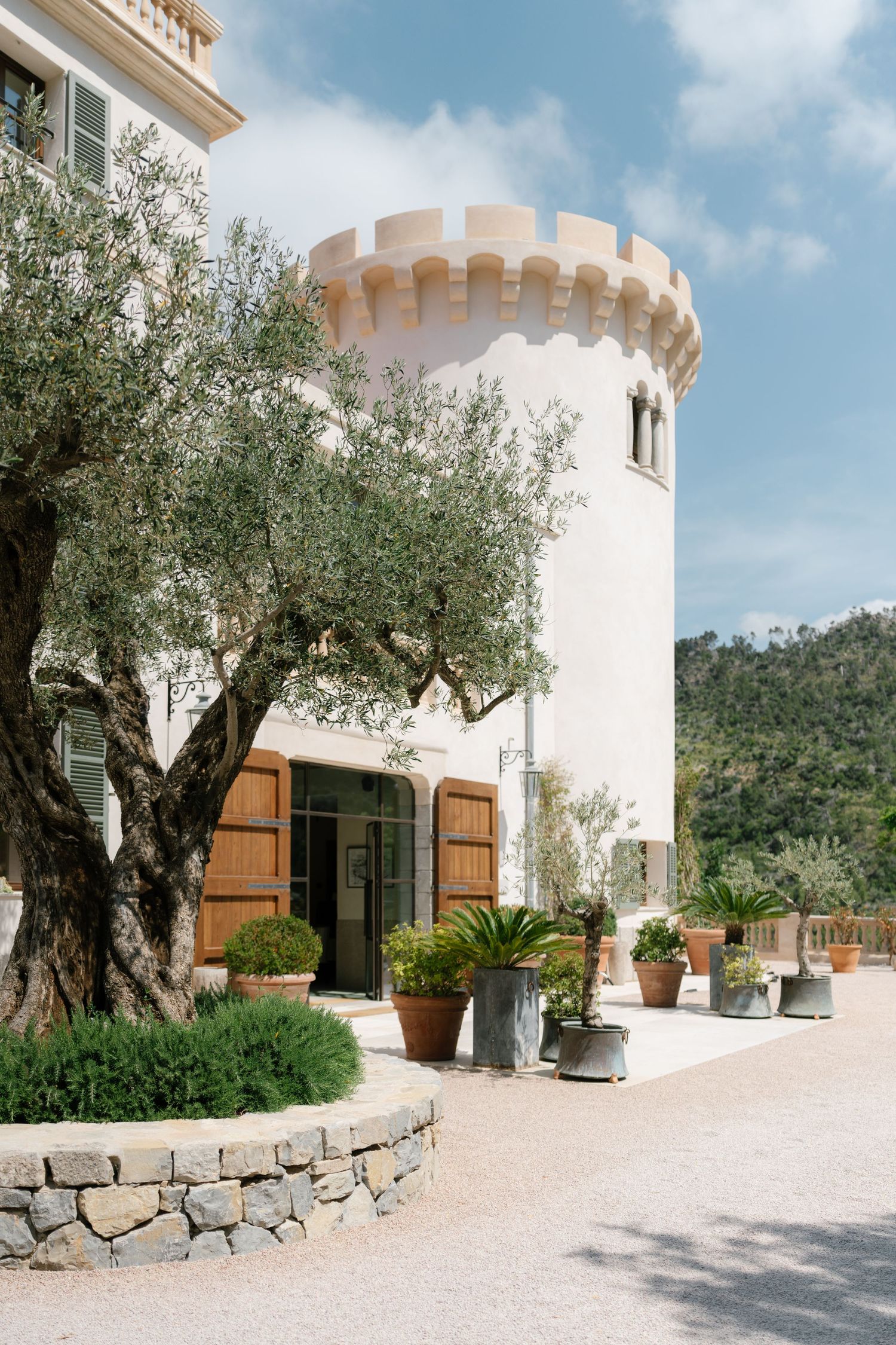 Mediterranean villa entrance with white tower, wooden shutters, olive trees and potted plants along stone pathway.