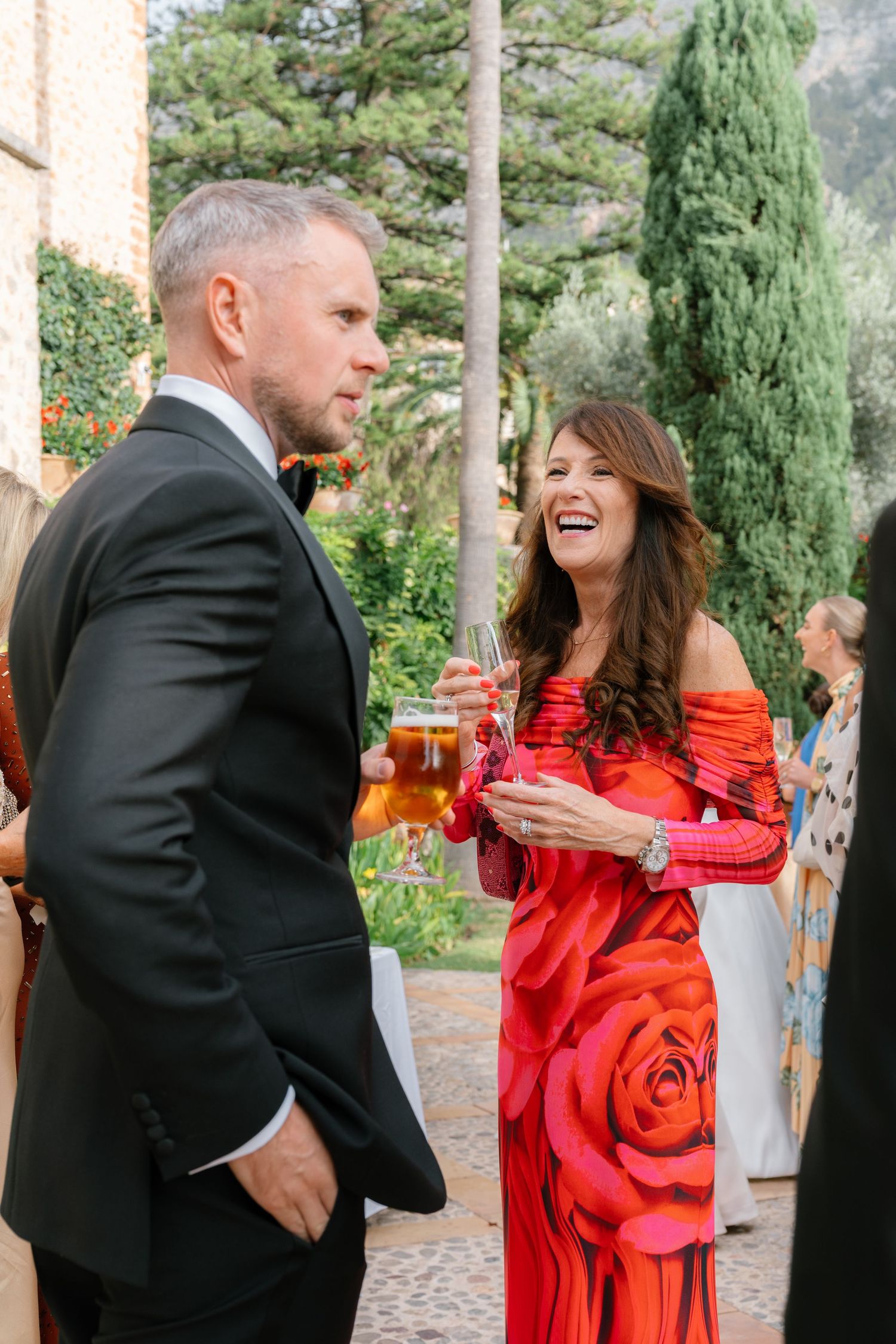 Wedding guests in formal attire share animated conversation at outdoor garden reception.