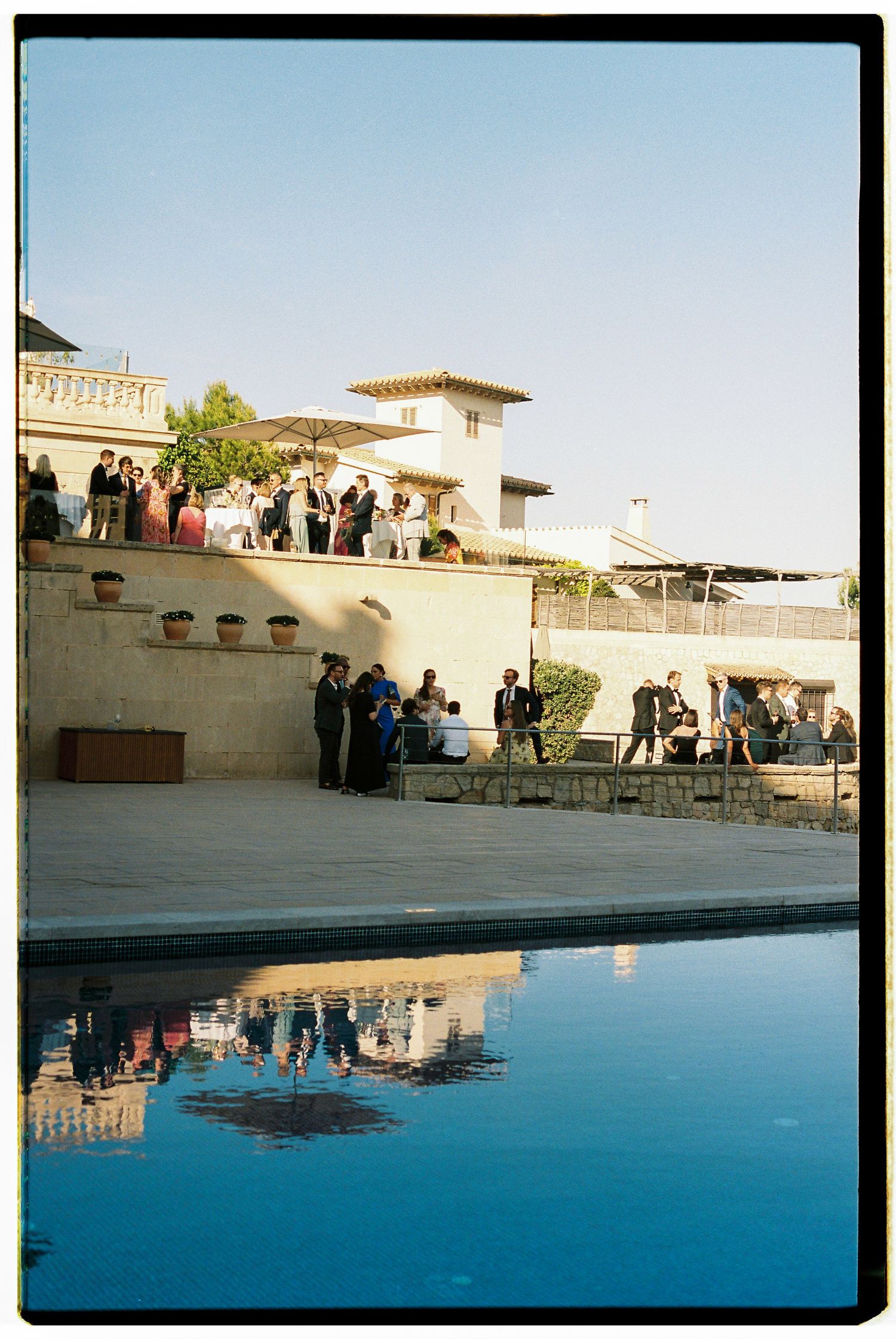 Guests gather around a luxurious swimming pool at an elegant venue with white architectural features.