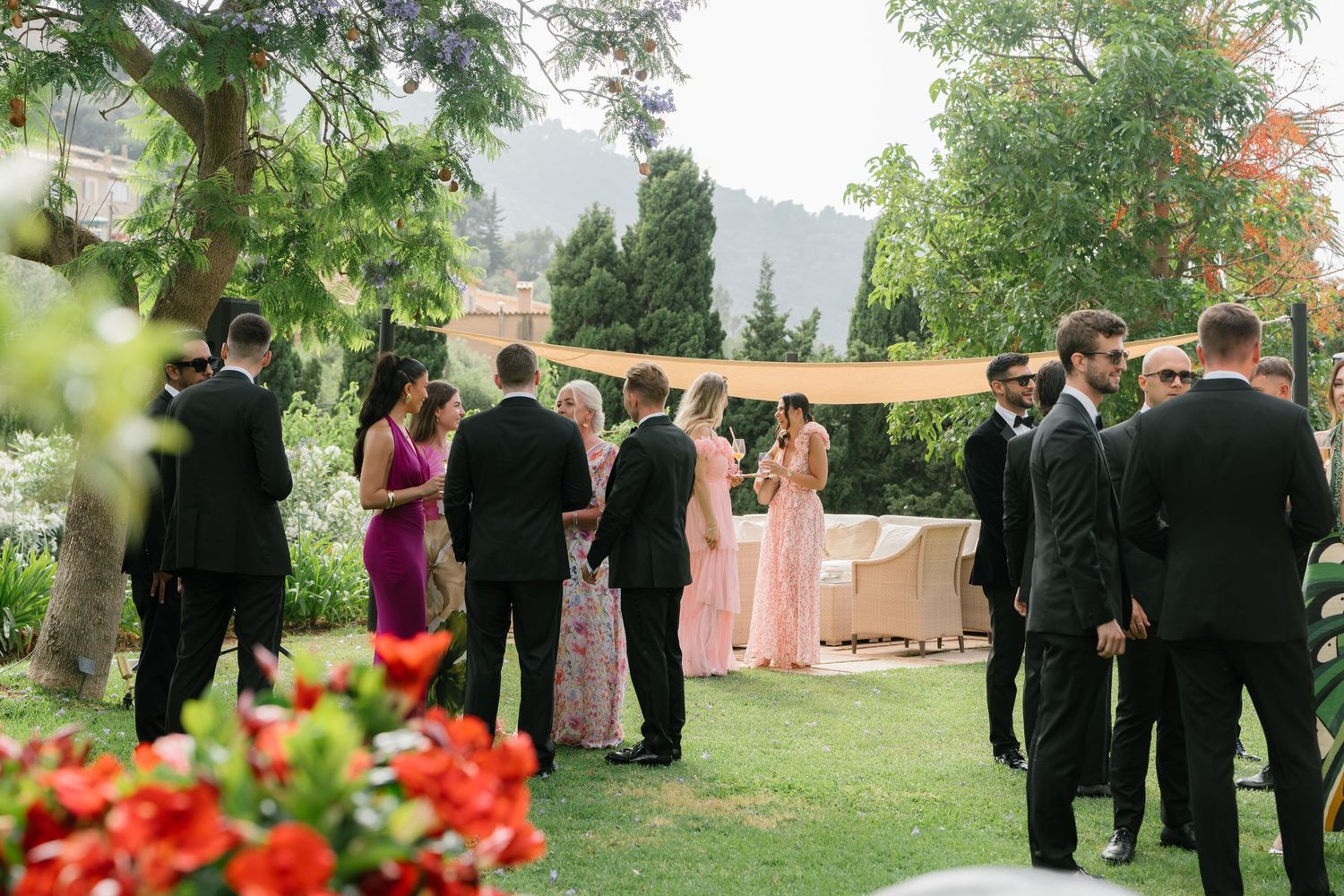 Outdoor wedding ceremony setup with guests gathered among trees and colorful flowers.