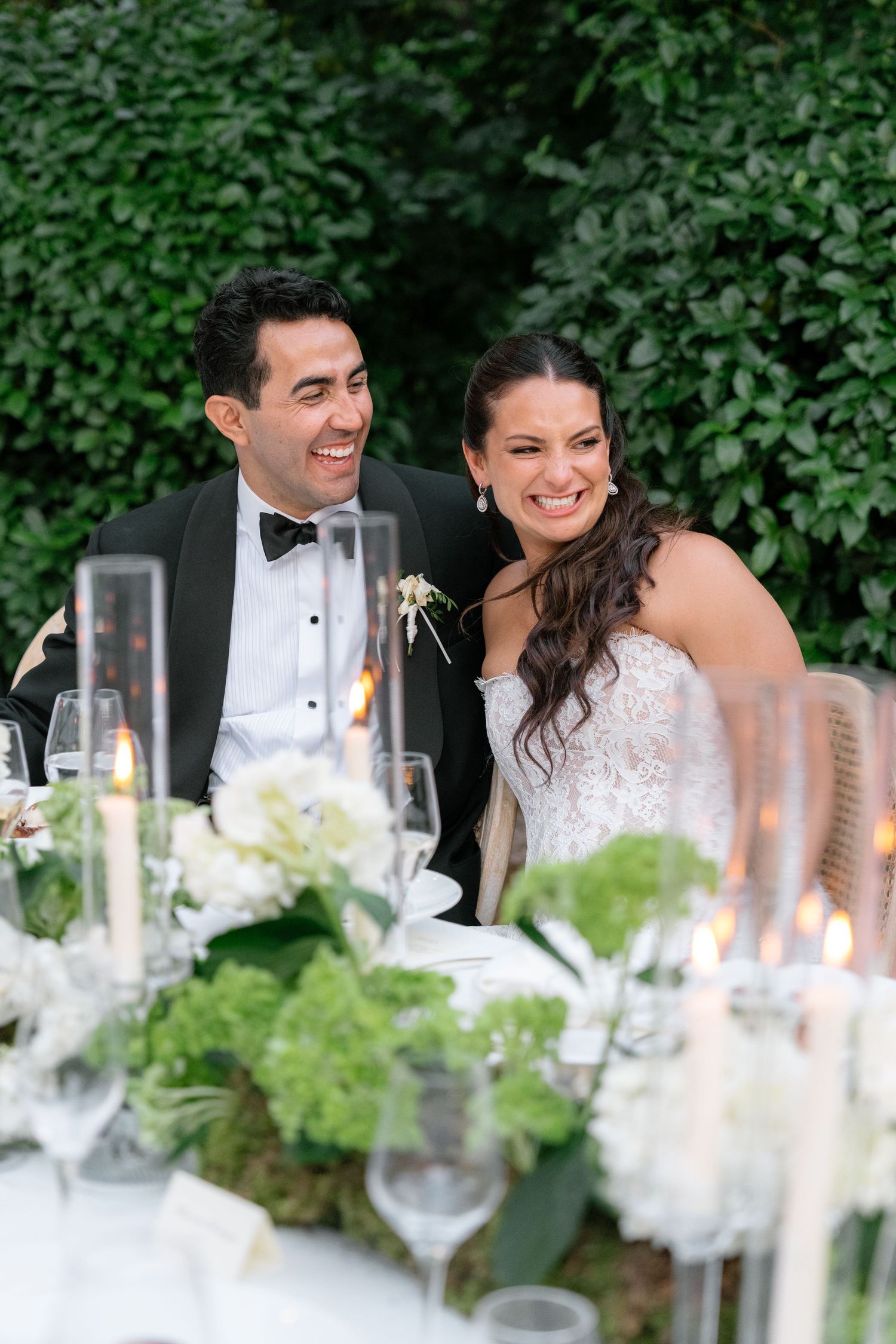 Smiling bride and groom seated at elegant wedding reception table with white floral arrangements and candles.