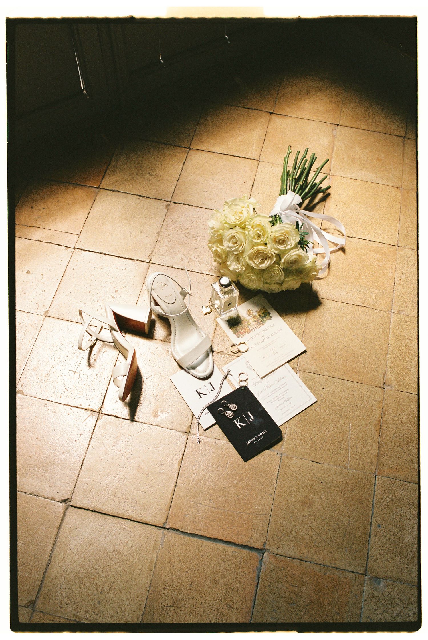 Bridal flat lay on terracotta tiles featuring white heels, rose bouquet, rings, and wedding stationery in warm natural light.