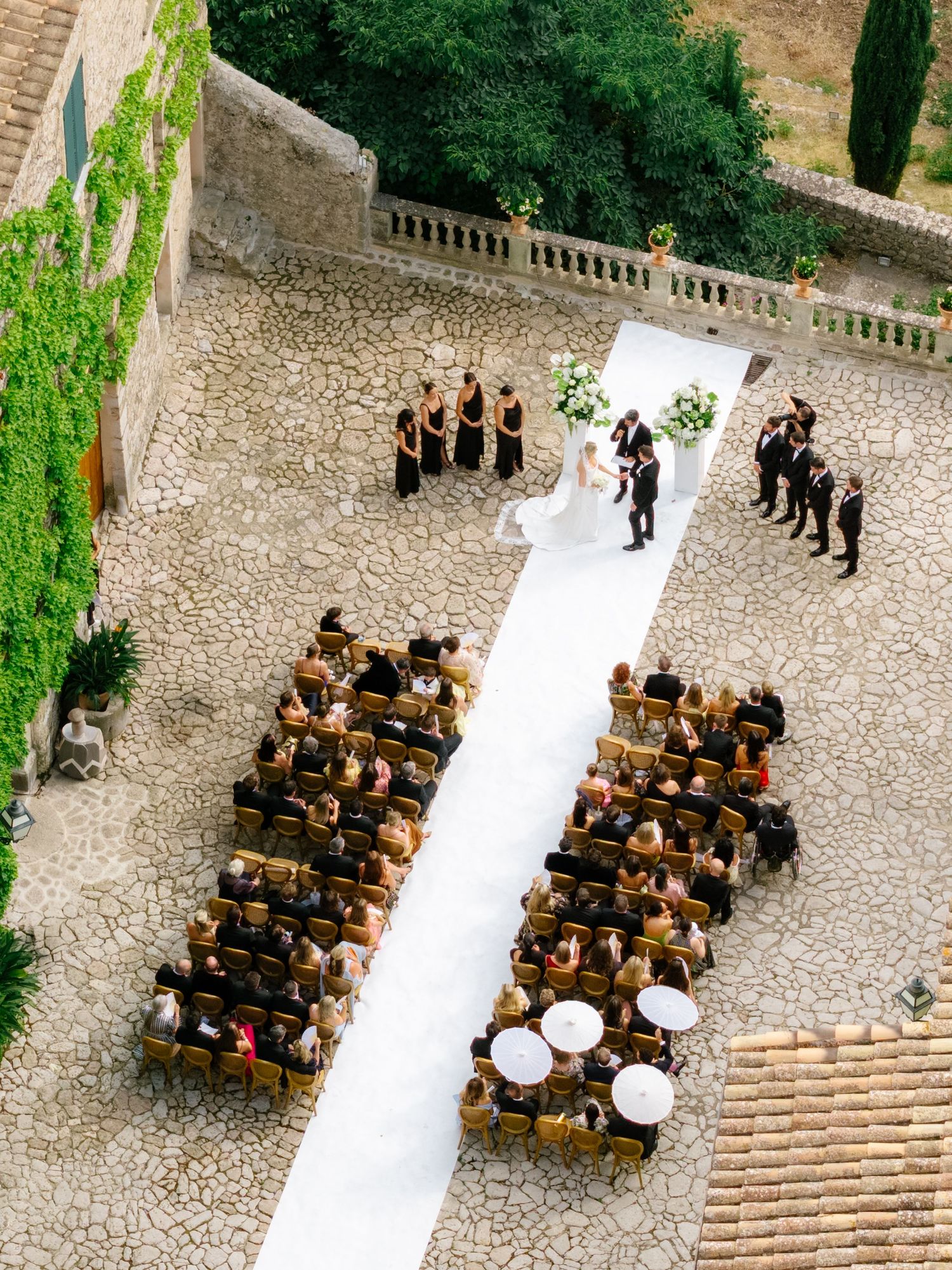 Aerial view of an elegant outdoor wedding ceremony with white aisle runner, seated guests, and bridal party at a stone courtyard venue.