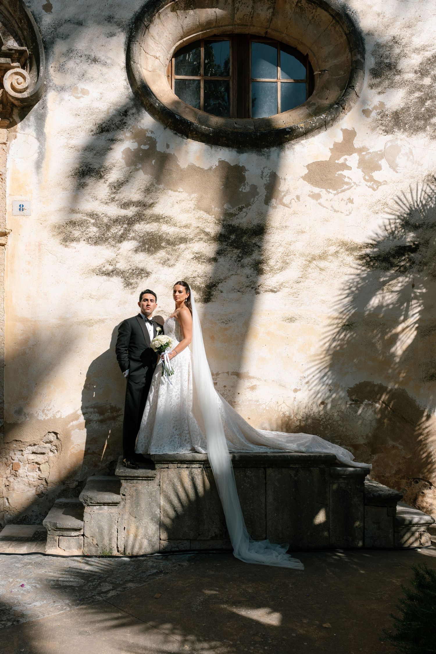 Bride and groom pose on stone steps against a rustic villa wall with a round window during their wedding.