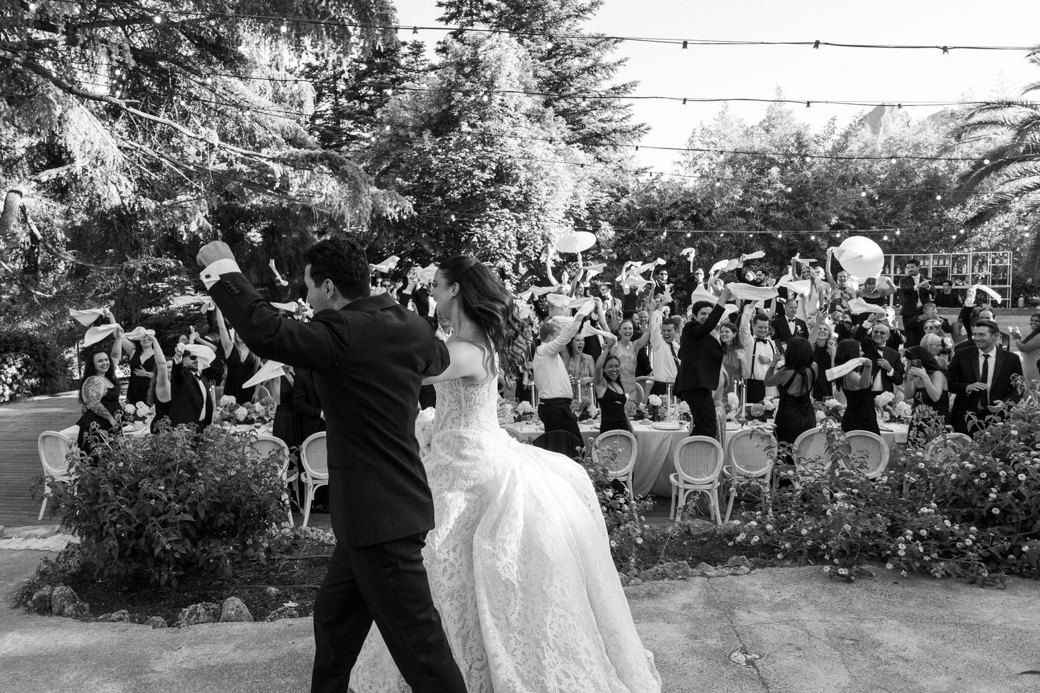 Black and white photo of bride and groom sharing first dance at outdoor wedding reception with guests celebrating.