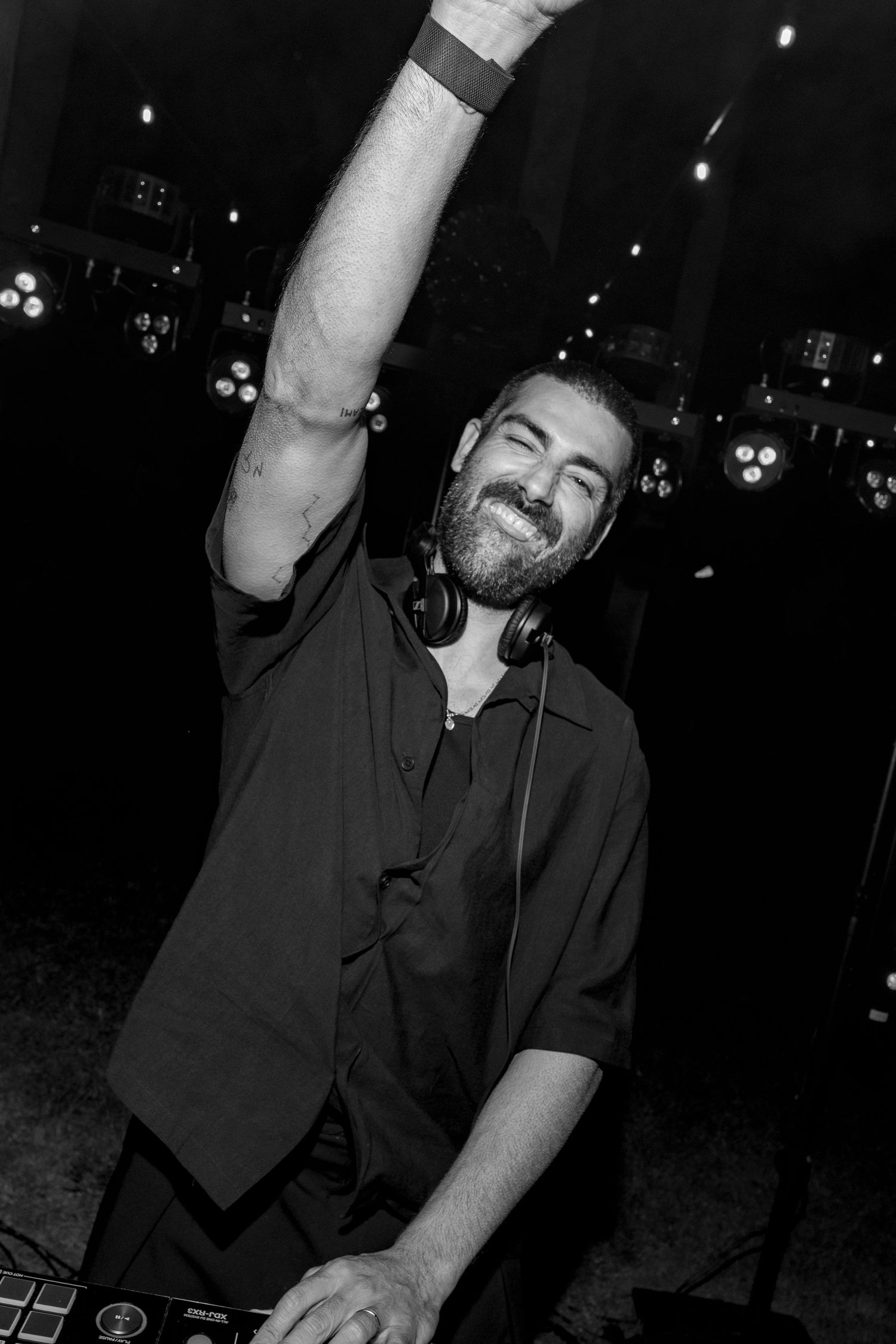A smiling bearded DJ raises one hand in celebration while wearing headphones around his neck at a nightclub event.