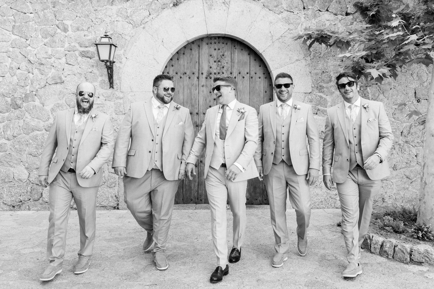 Groomsmen in light beige suits posing against a stone wall in black and white photography.