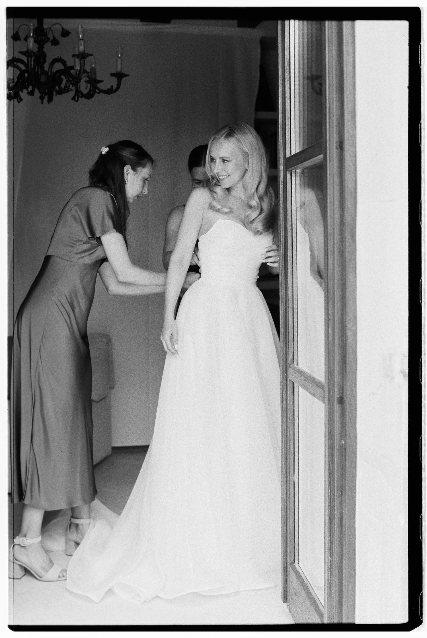 Black and white wedding preparation photos showing bride in white dress and bridesmaids helping with final touches.