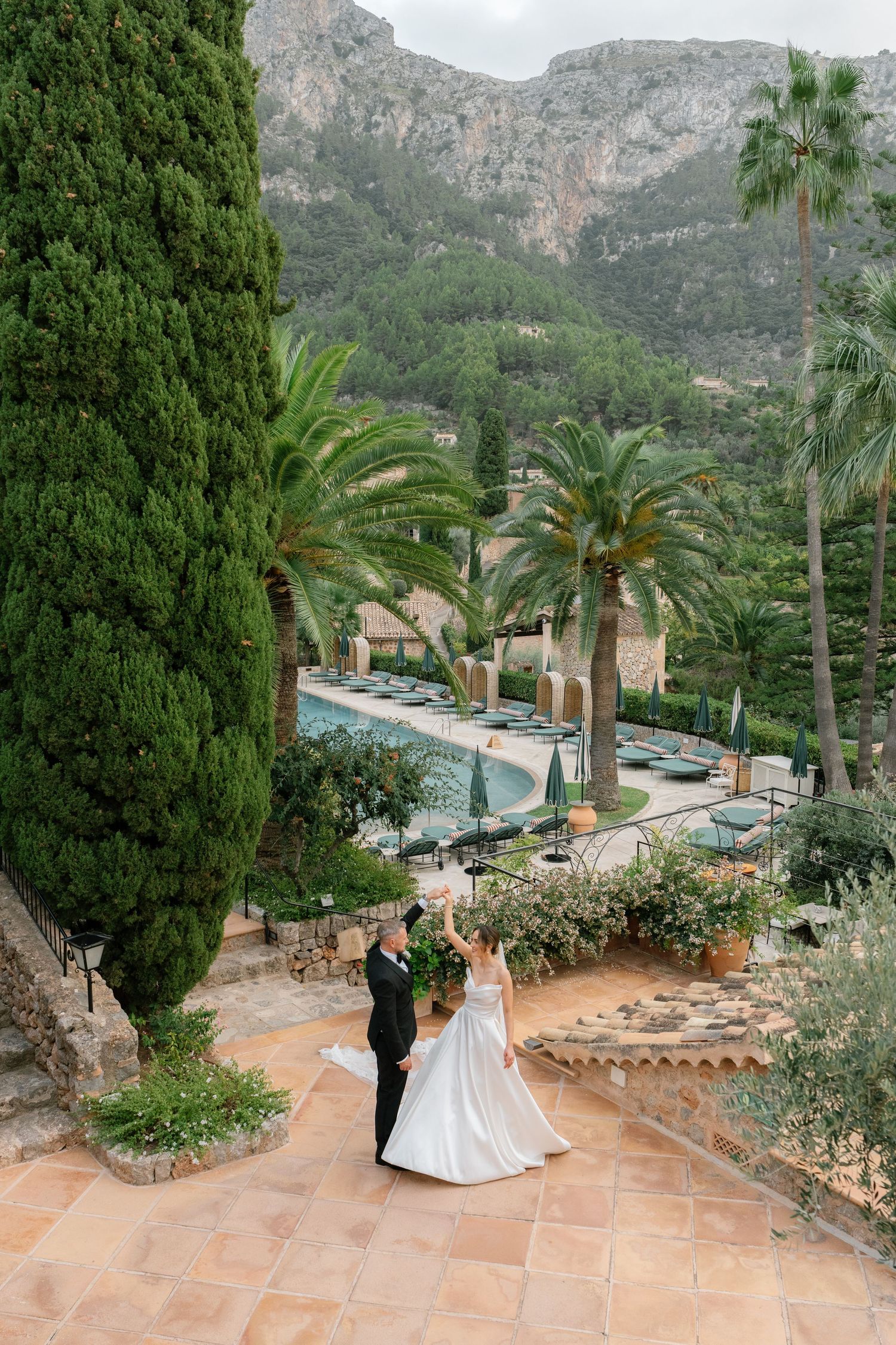 A scenic wedding venue with mountain views features a couple on a tiled terrace with palm trees.