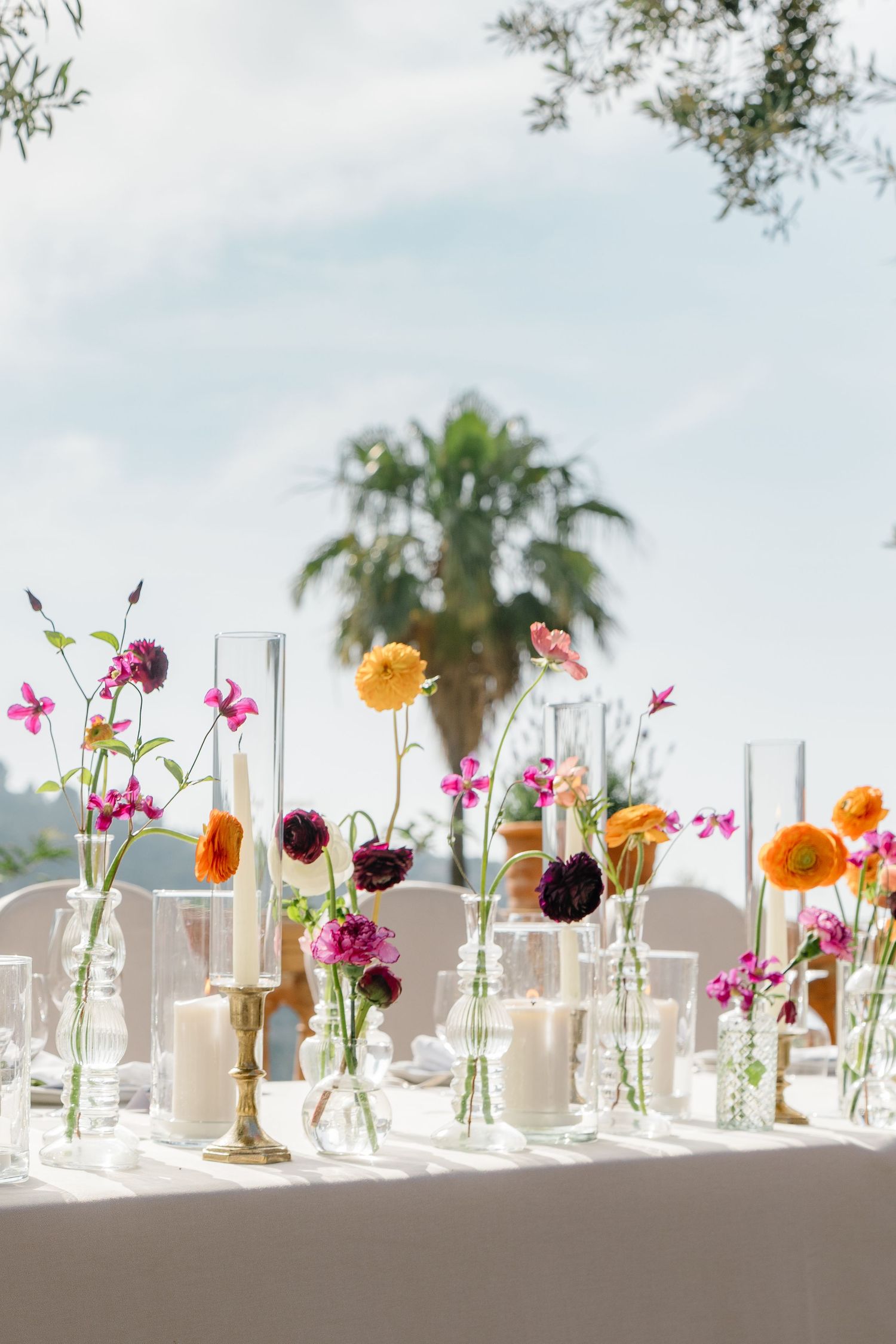 Wedding reception table decorated with colorful spring flowers in glass vases against blue sky.