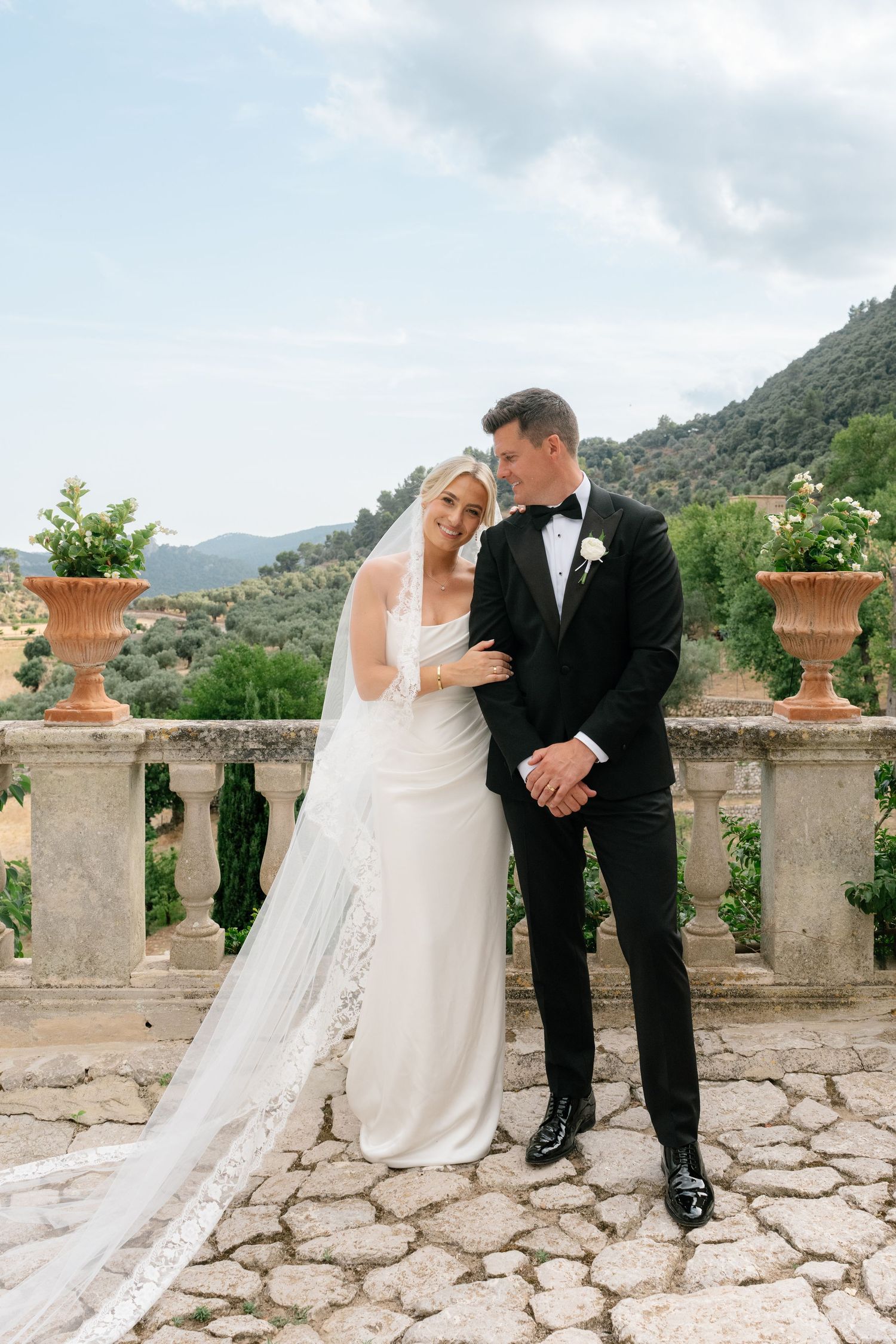 Bride in white gown and veil with groom in black tuxedo posing on stone terrace with scenic mountain views.