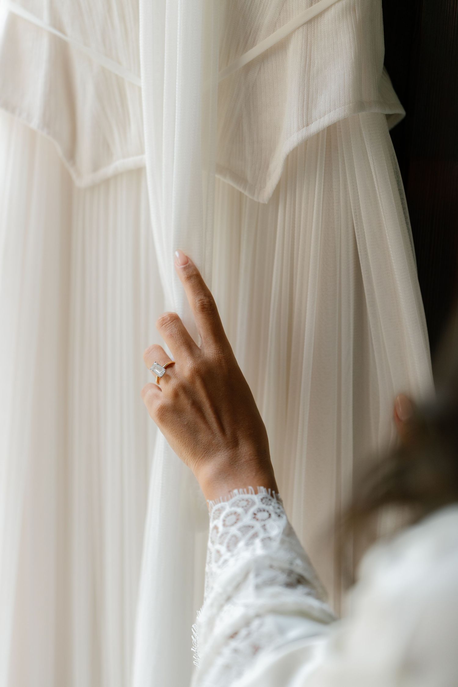 A hand wearing a lace sleeve and engagement ring touches a flowing white wedding dress hanging on display.